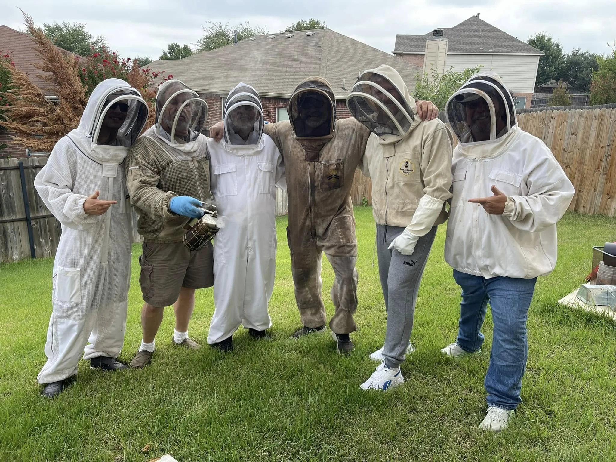 Group of six people wearing beekeeping suits with veiled hats standing in a backyard with a wooden fence, green grass, trees, and houses in the background.