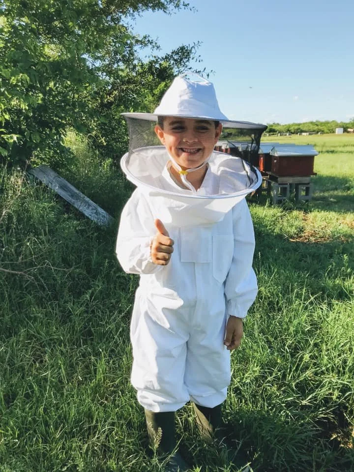 A young boy dressed in a white beekeeping suit and hat with a veil, standing outdoors in a grassy area, smiling and giving a thumbs up.