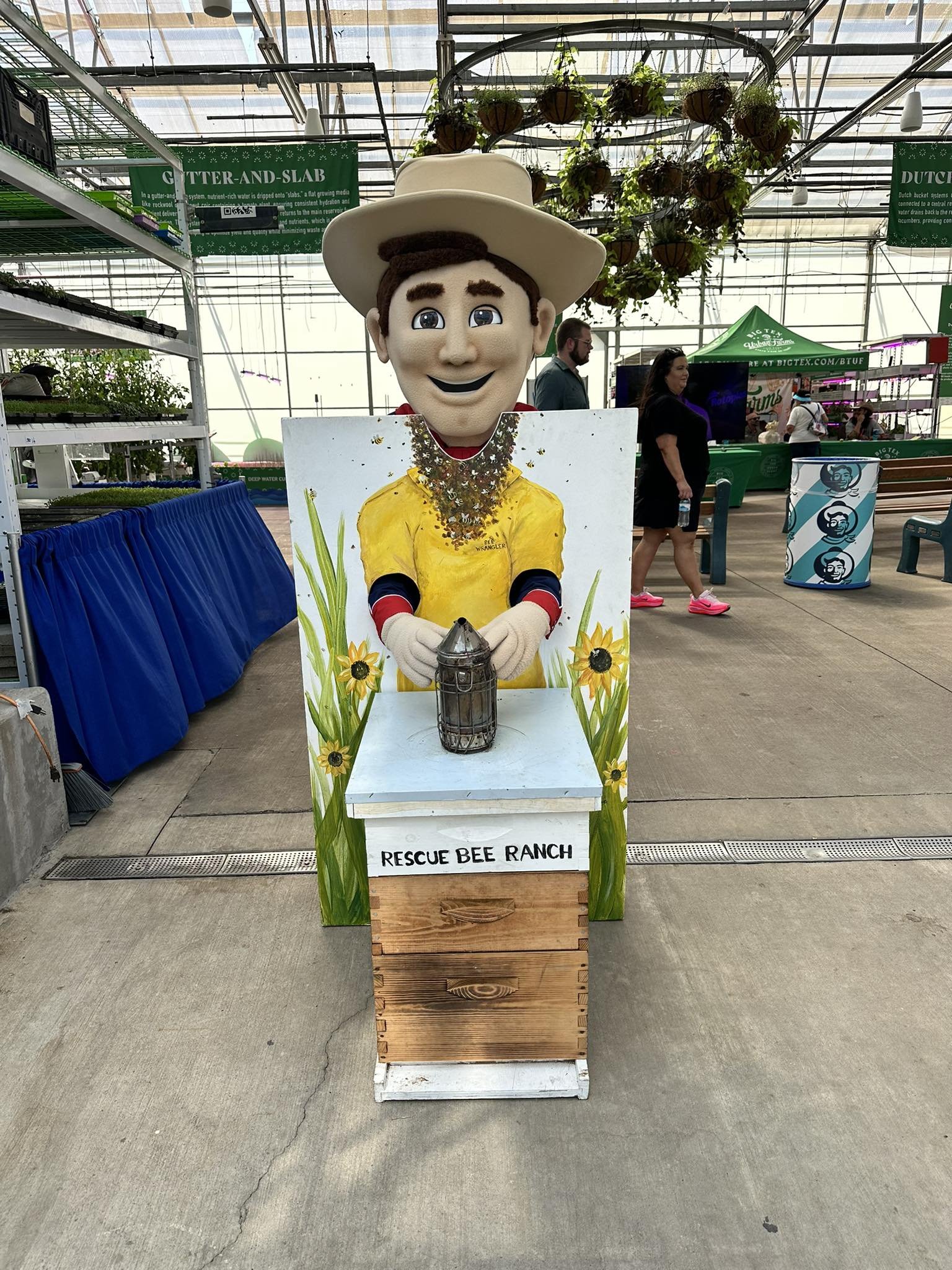 A person in a bee costume stands behind a painted sign reading "Rescue Bee Ranch" inside a large greenhouse or conservatory with other visitors and displays in the background.