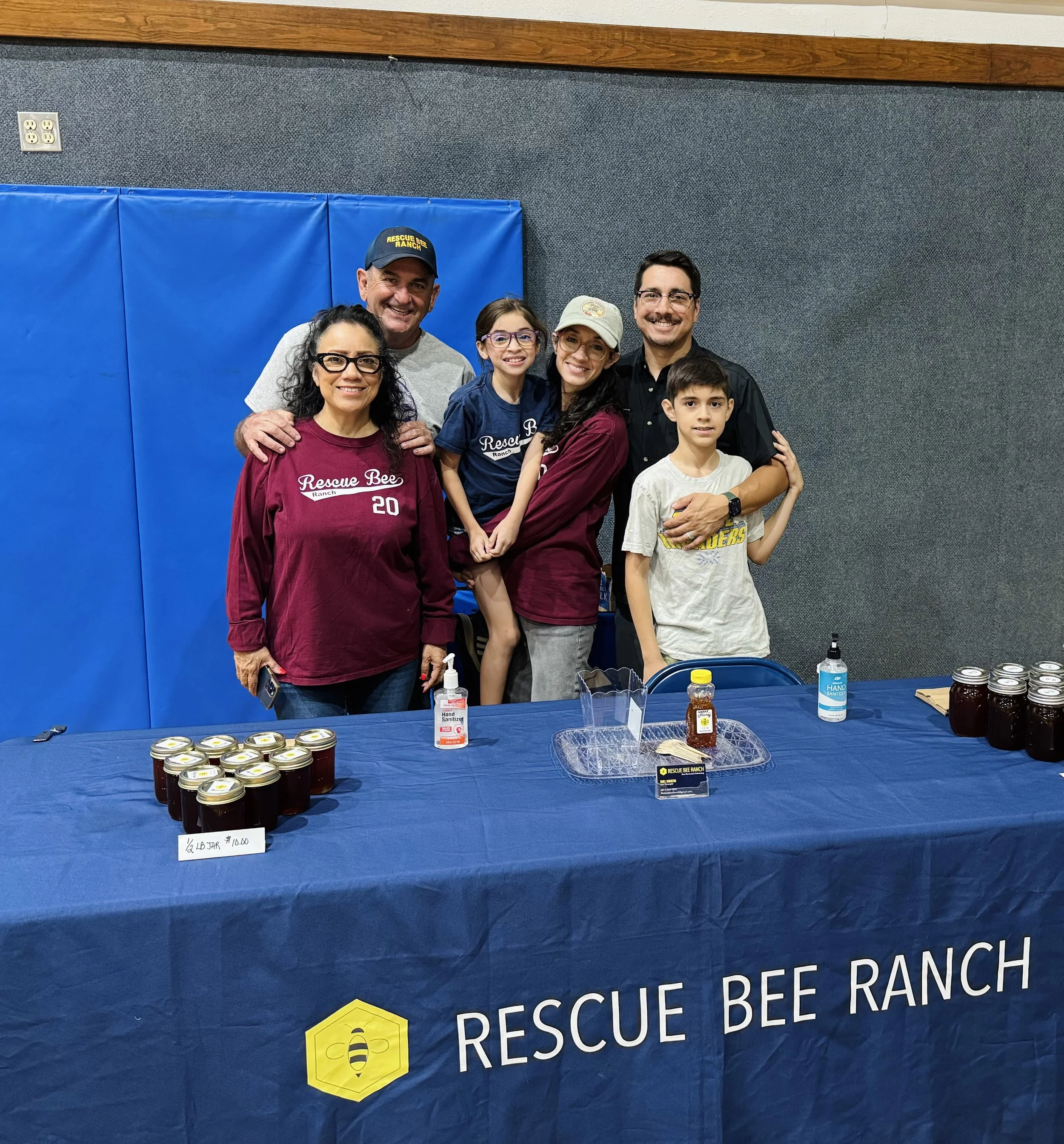 Group of six people, including children and adults, gathered at a booth with a blue tablecloth displaying jars of honey, hand sanitizer, and honey tasting samples in a community or event setting. The background features a blue padded wall and a sign that reads "Rescue Bee Ranch." The group is smiling and posing for the photo.