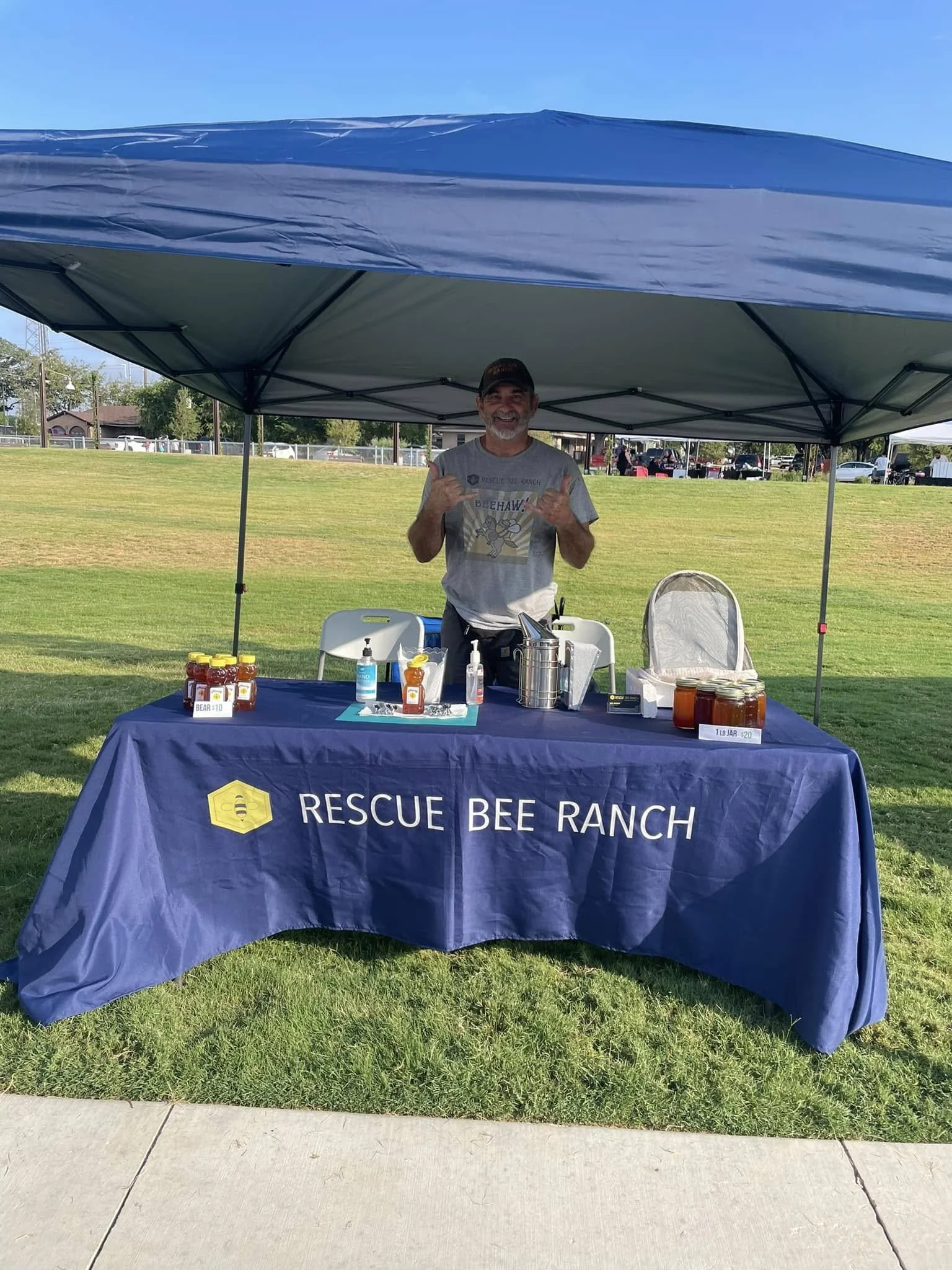 A man standing behind a table under a canopy at an outdoor event, with various bottles and jars on the table, and a banner that reads 'Rescue Bee Ranch'.