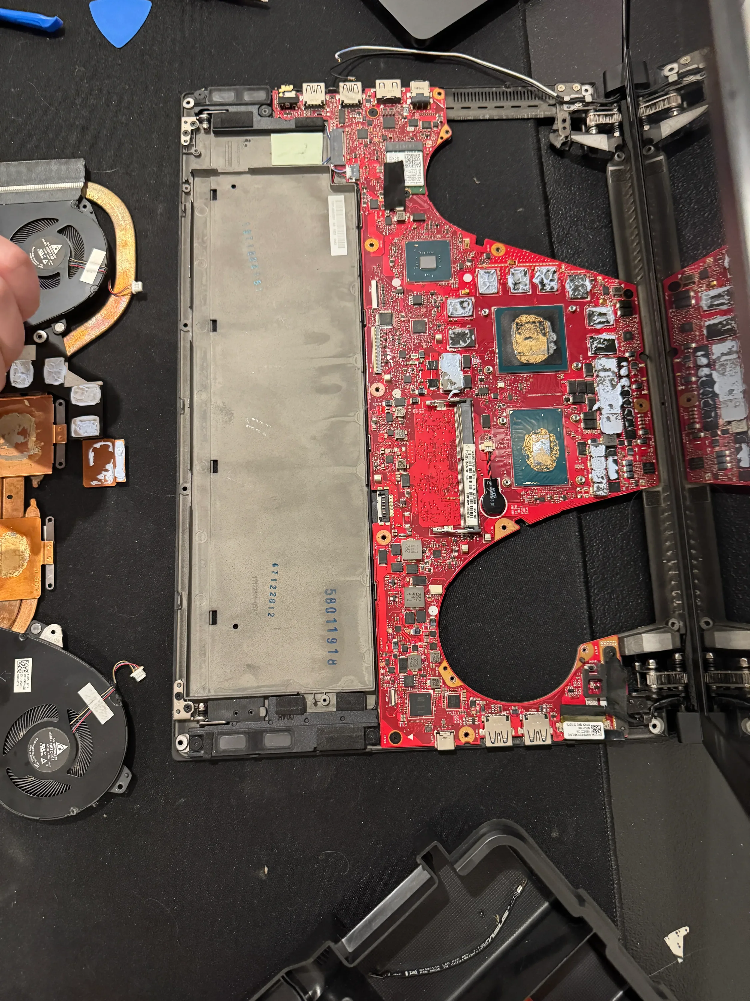 A partially disassembled laptop with its motherboard exposed. The main red circuit board with various chips and components is visible, along with cooling fans to the left. Tools are on the black work surface.