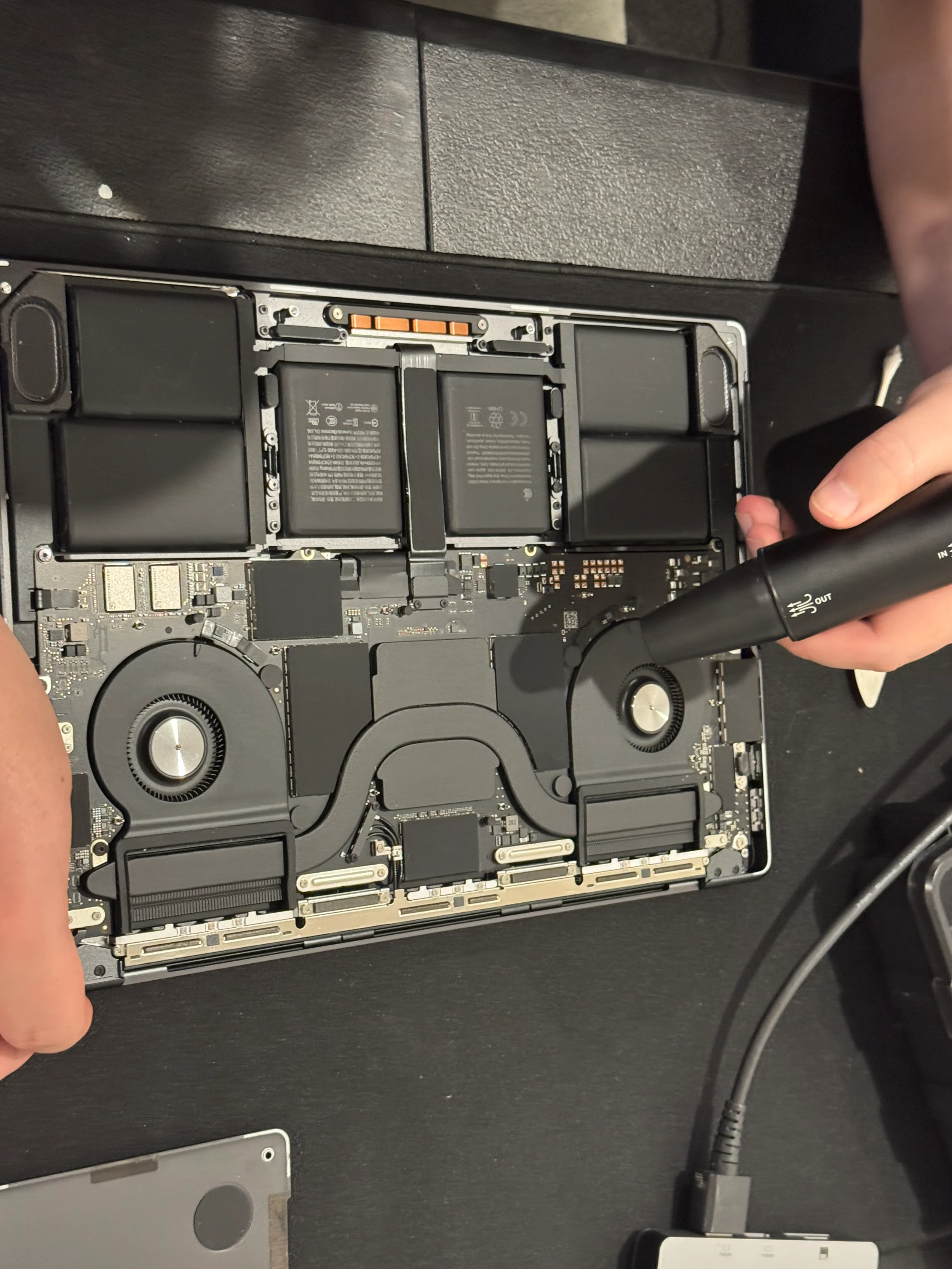 An opened electronic device, possibly a laptop or tablet, on a black surface with a person's hand holding a small vacuum or air blower cleaning the interior components, including batteries and circuit boards.