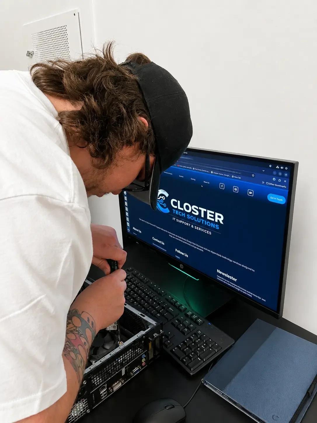 Person working on a computer with the screen displaying Closter Tech Solutions website, wearing glasses and a backwards baseball cap.