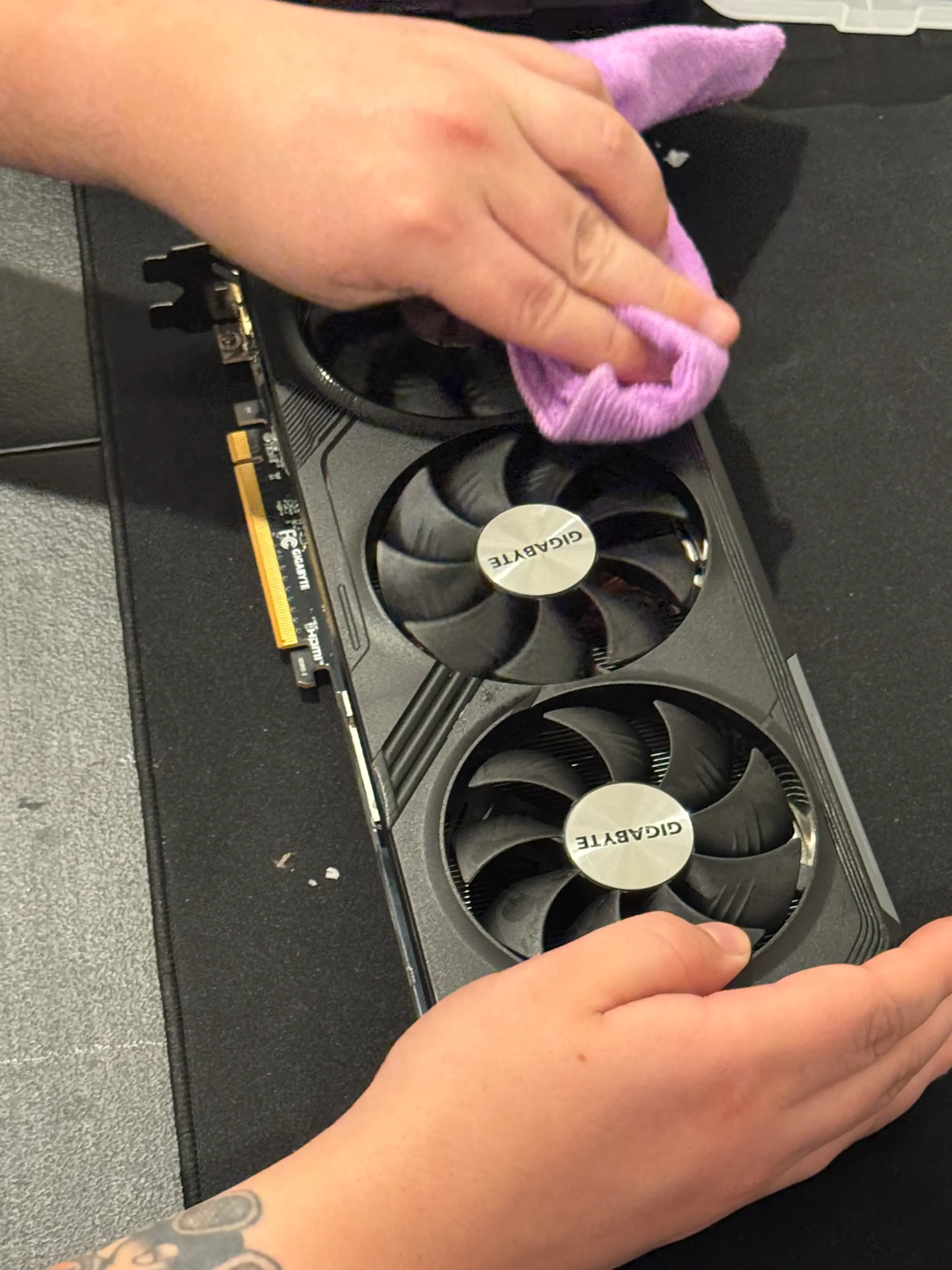 A person's hand is wiping a graphics card with a pink cloth. The graphics card is on a black surface and has two fans with the word 'GIGABYTE' on the center of one fan.