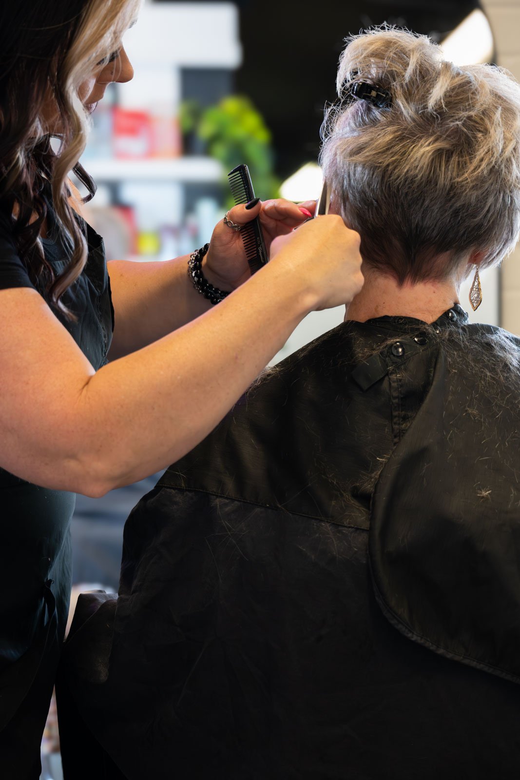 A hairstylist cutting a woman's hair in a salon, with the stylist holding scissors and a comb, and the woman sitting with a black cape around her shoulders.