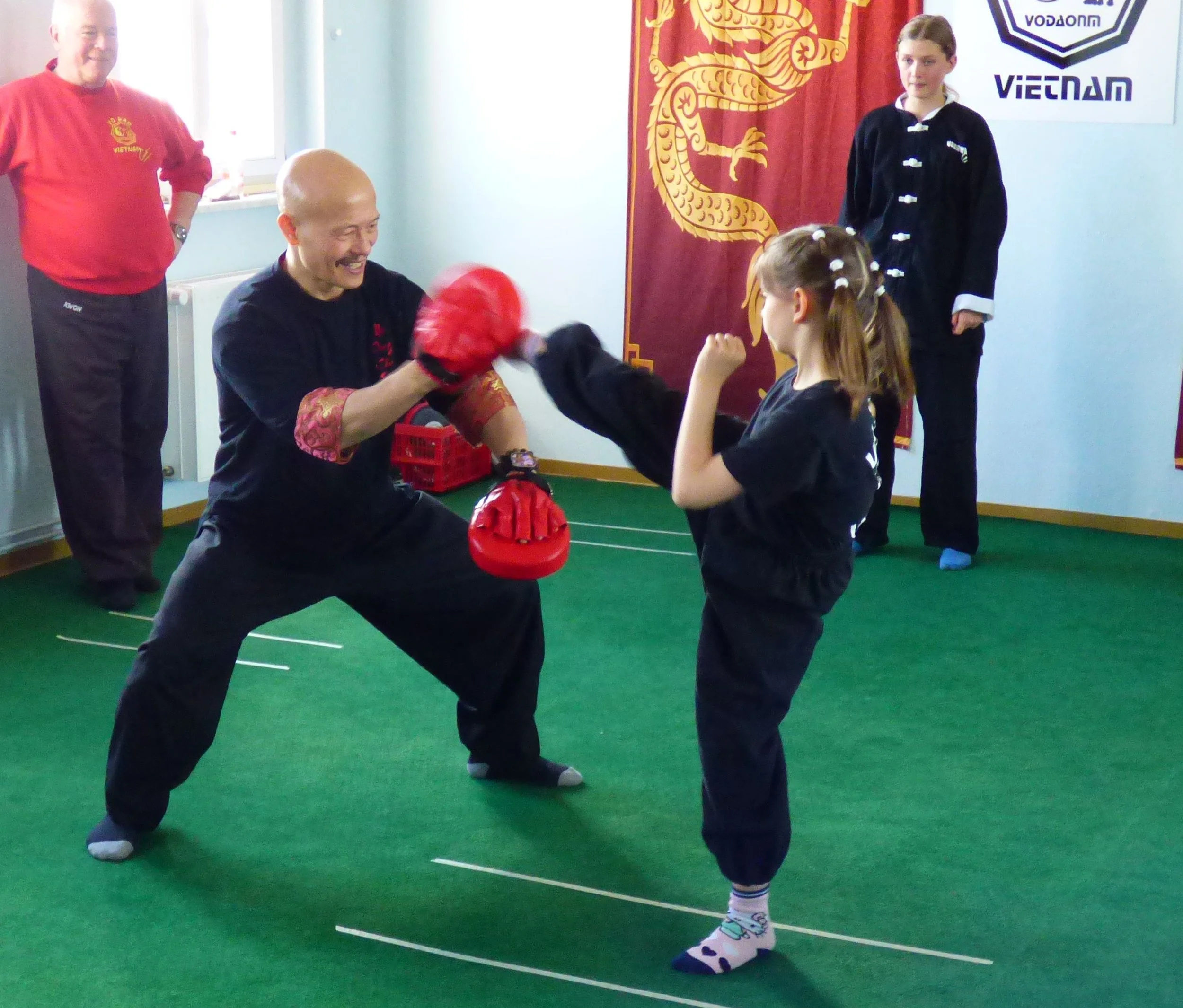 Zwei Kinder beim Karate-Training, eine Person trägt einen schwarzen Anzug, eine andere Person hat eine rote Boxhandschuh, im Hintergrund eine Fahne mit Löwe und ein Logo mit der Aufschrift "Vietnam".