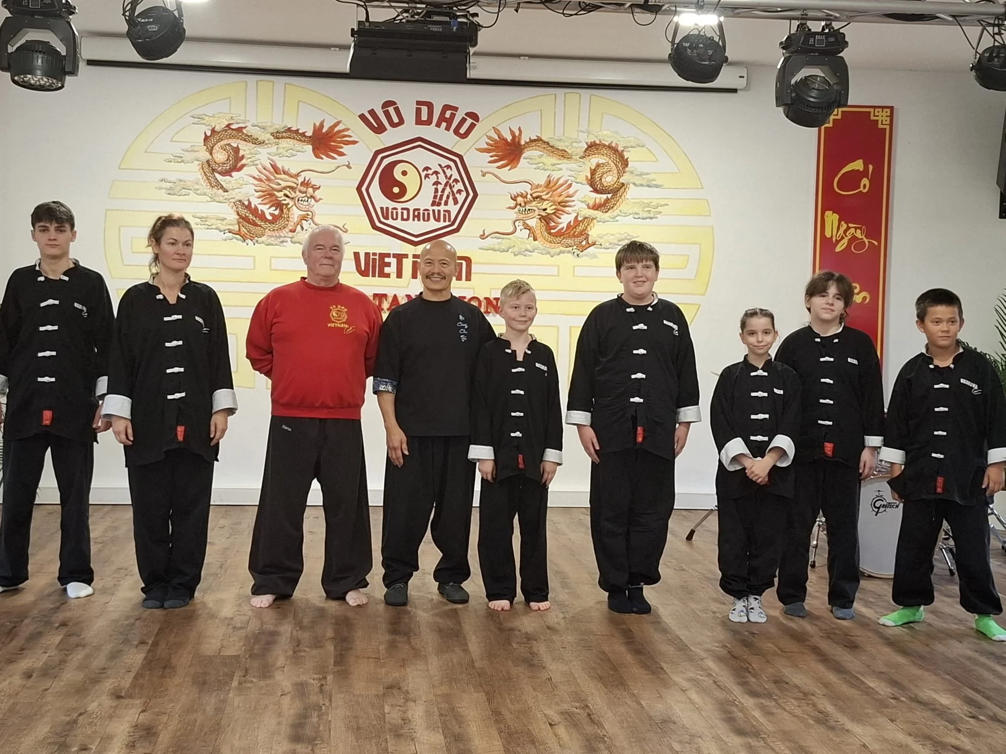 Gruppe von Menschen in schwarzen Uniformen, einige mit weißen Schuhen, steht in einer Reihe auf einem Holzfußboden. Zwei Männer in der Mitte tragen rote Jacken. Hintergrund zeigt eine Wand mit einem großen chinesischen Drachen und asiatischer Schrift.