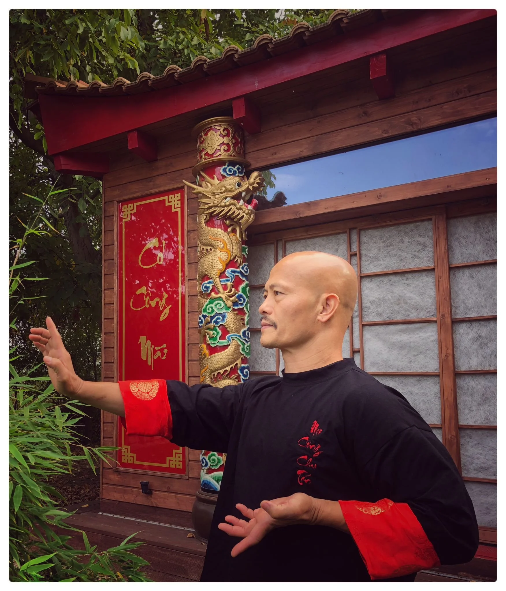 Ein Mann in traditioneller chinesischer Kleidung führt eine Tai-Chi-Übung im Freien vor, vor einem Holzzaun mit einem roten Banner und einem dekorativen chinesischen Drachen-Säulenstück. Es sind Bäume im Hintergrund.