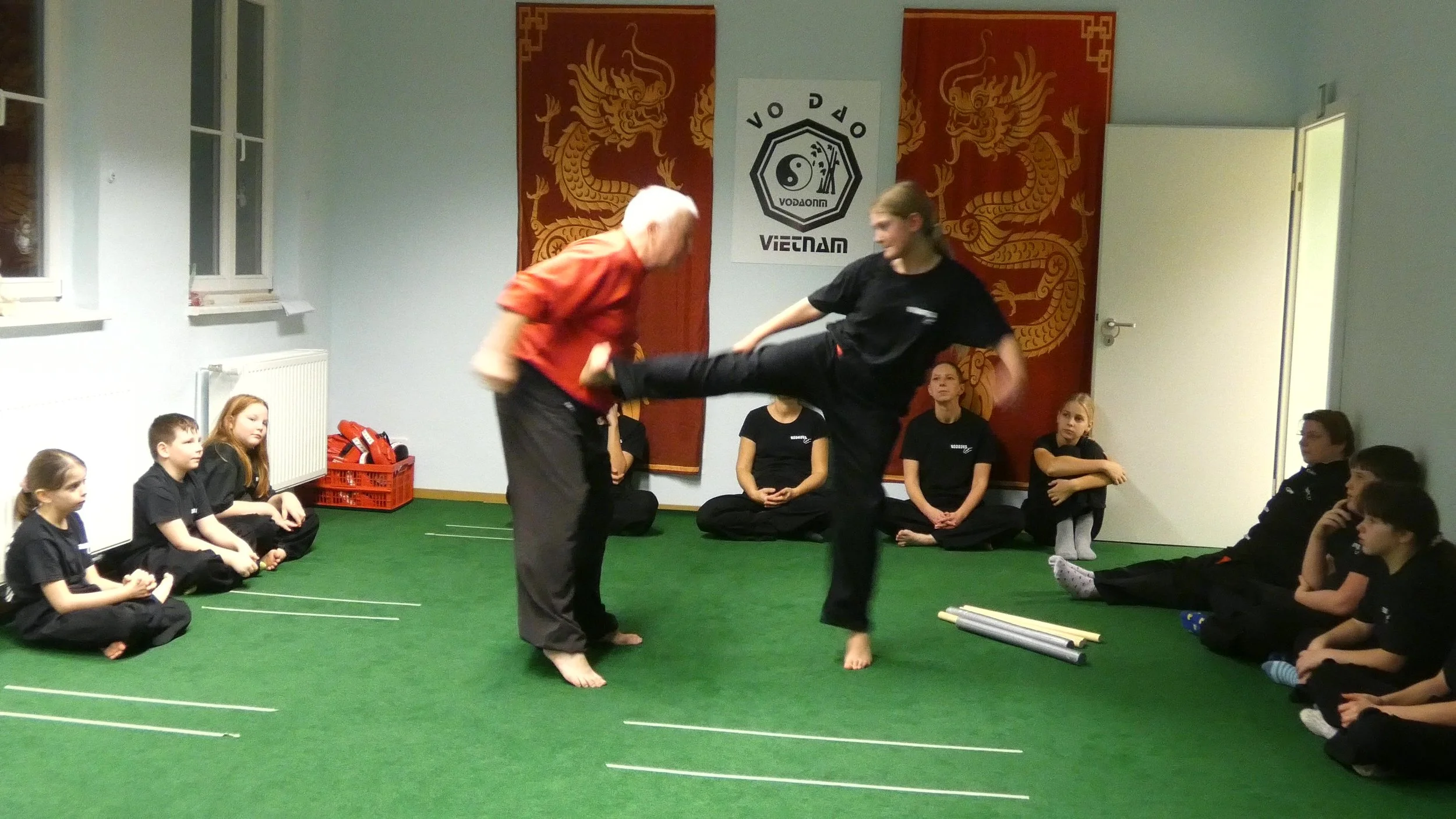 Martial Arts Unterricht mit älterem Mann und jüngerer Frau in einem Raum mit grüner Matte und chinesischen Drachentapeten an der Wand, die Kinder sitzen im Kreis auf dem Boden und beobachten, während die Frau einen Kick gegen den Mann ausführt.