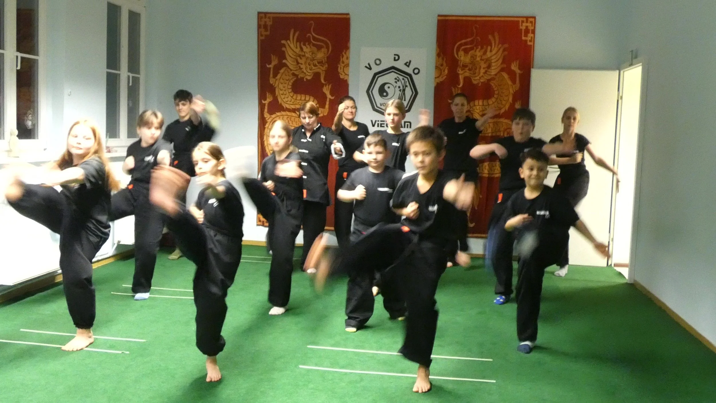 Gruppe junger Schüler in schwarzen Trainingsanzügen machen Kampfsportübungen in einem Raum mit grüner Matte, Backsteinlime und asiatisch inspirierten Wanddekorationen.