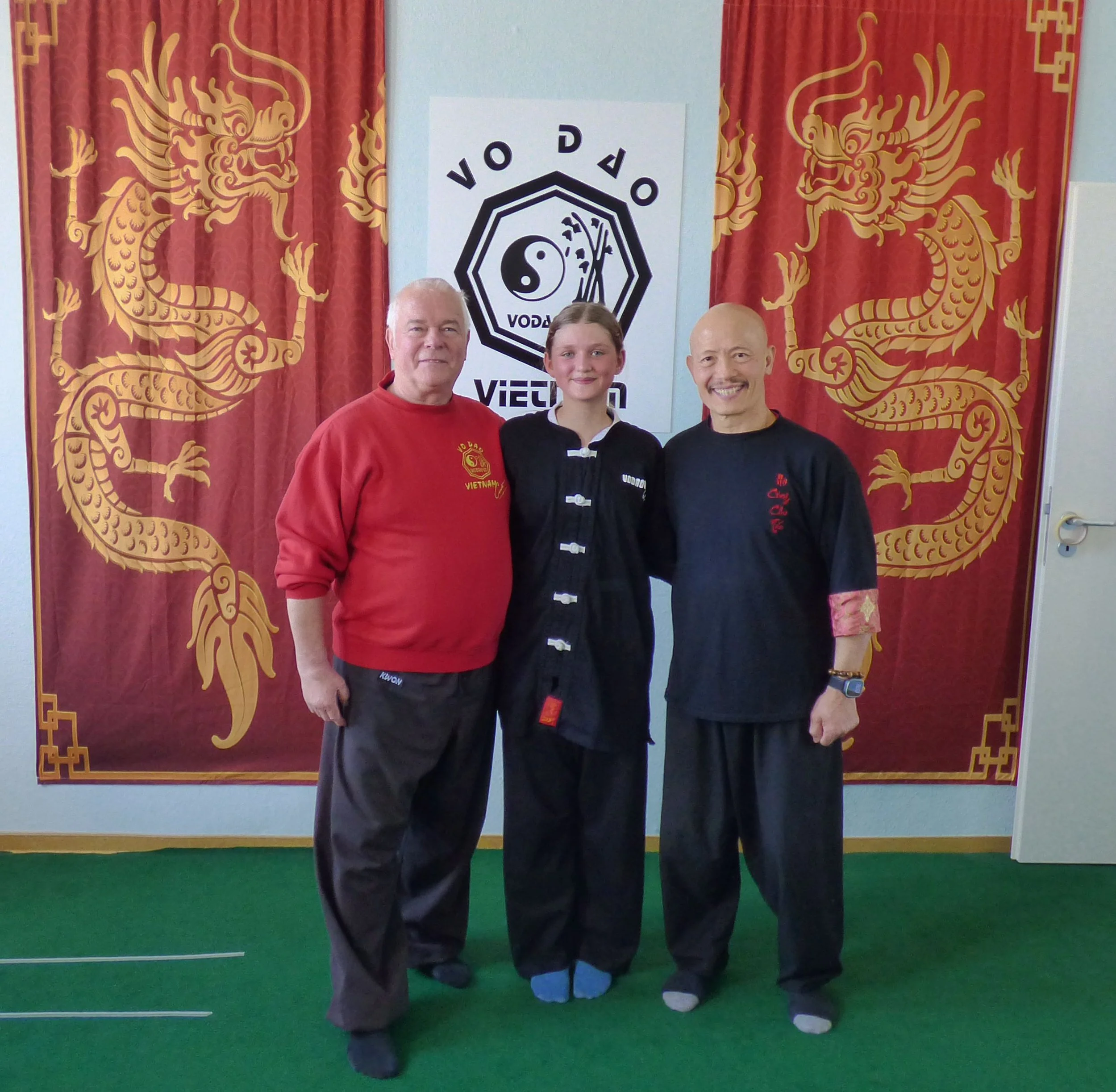 Drei Menschen in Schwarz und Rot vor einer chinesischen Drachentapete und einer Vovinam-Flagge, die Yin-Yang-Symbol und militärische Ausrüstung zeigt.