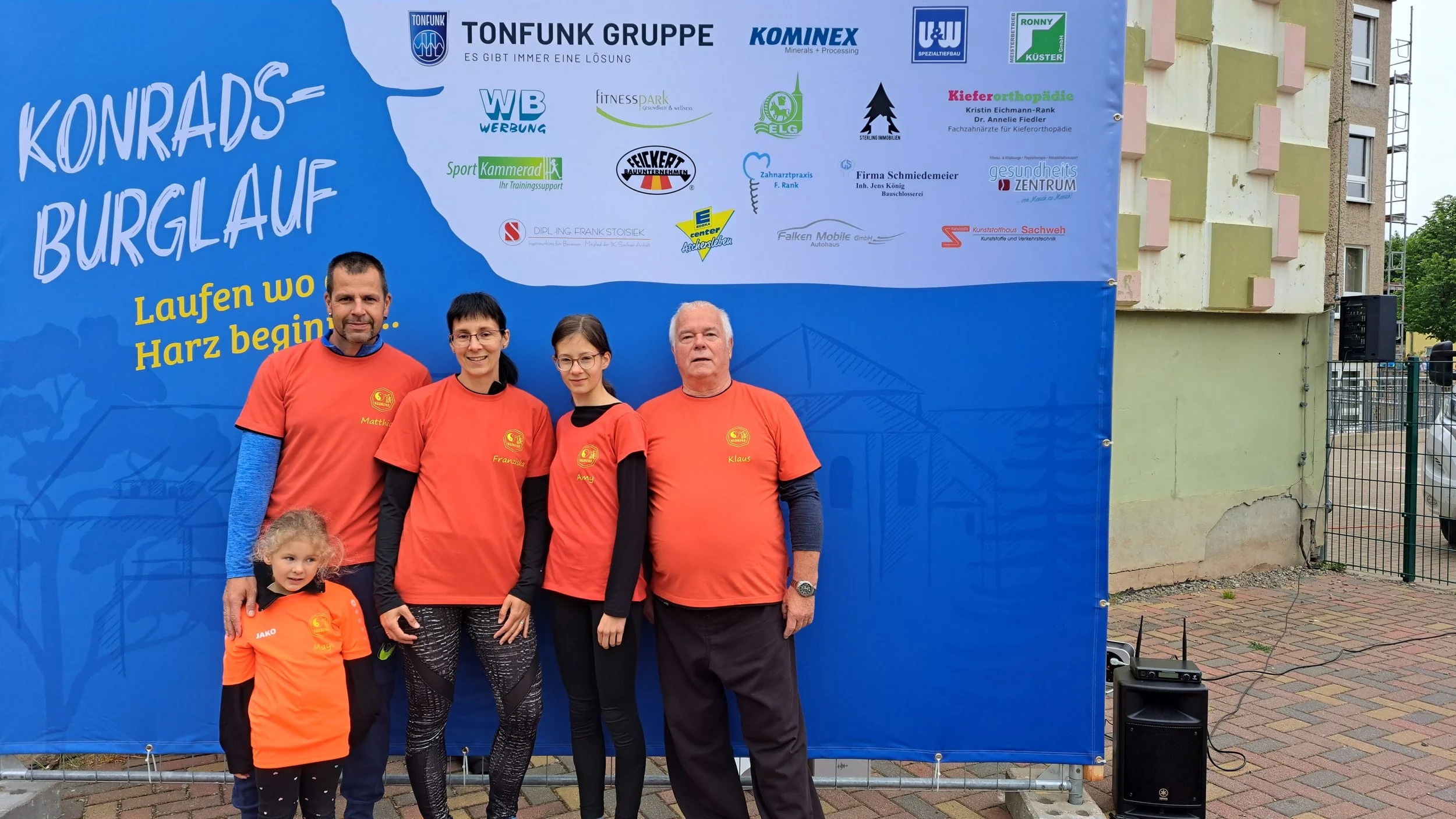 Gruppenbild bei einem Burglauf-Event, bestehend aus drei Erwachsenen und einem Kind, alle in orange T-Shirts mit Namen und einem Logo, vor einem großen blauen Banner mit Sponsorenlogos und dem Text "Konrads Burgauf", auf einer gepflasterten Straße, im Hintergrund ist ein teilweise gebautes Gebäude und ein Lautsprecher zu sehen.