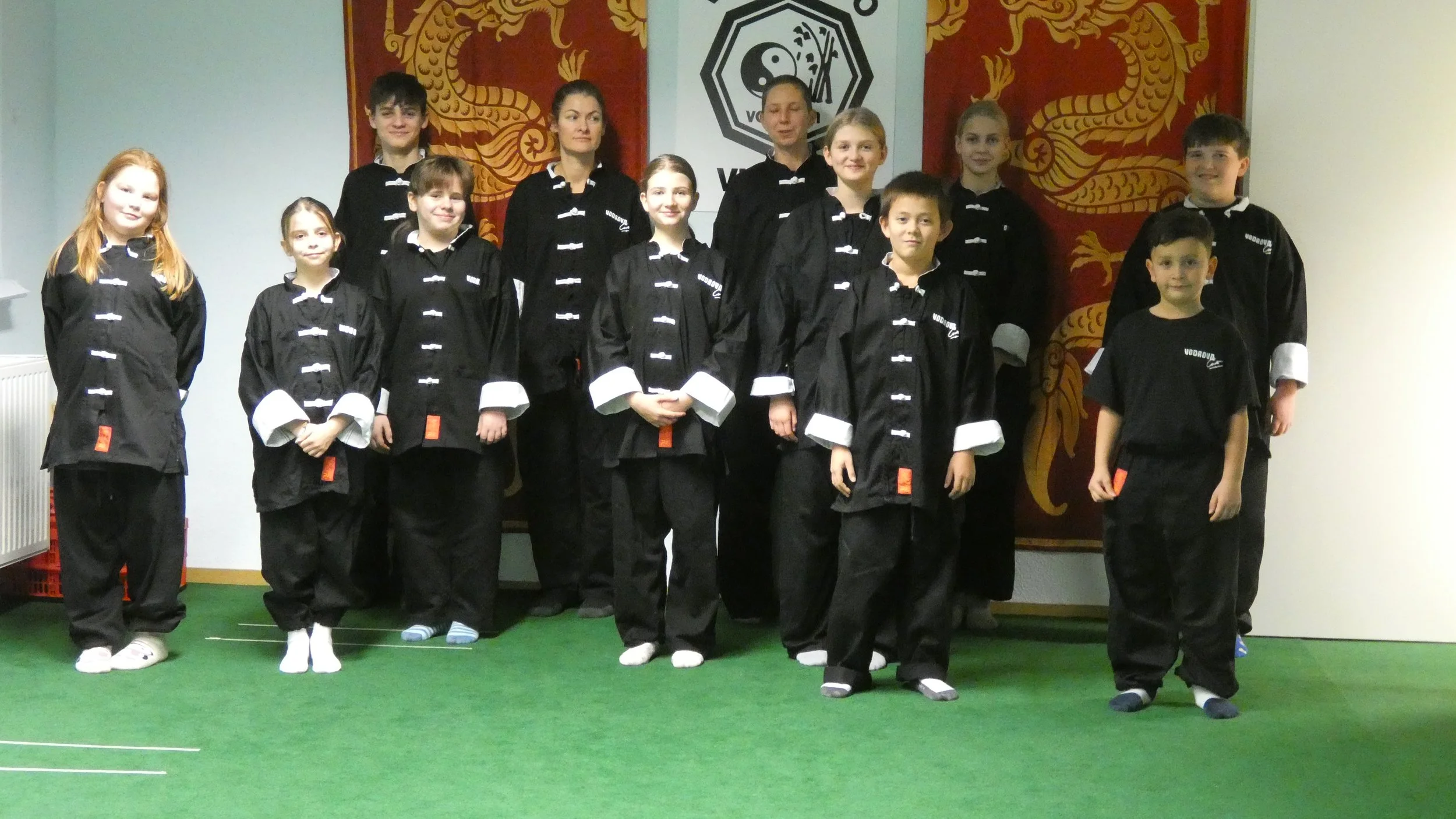 Gruppe von Kindern und Erwachsenen in schwarzen Kampfsportuniformen, die vor einer Wand mit chinesischen Drachendekorationen und einem Yin-Yang-Symbol stehen.