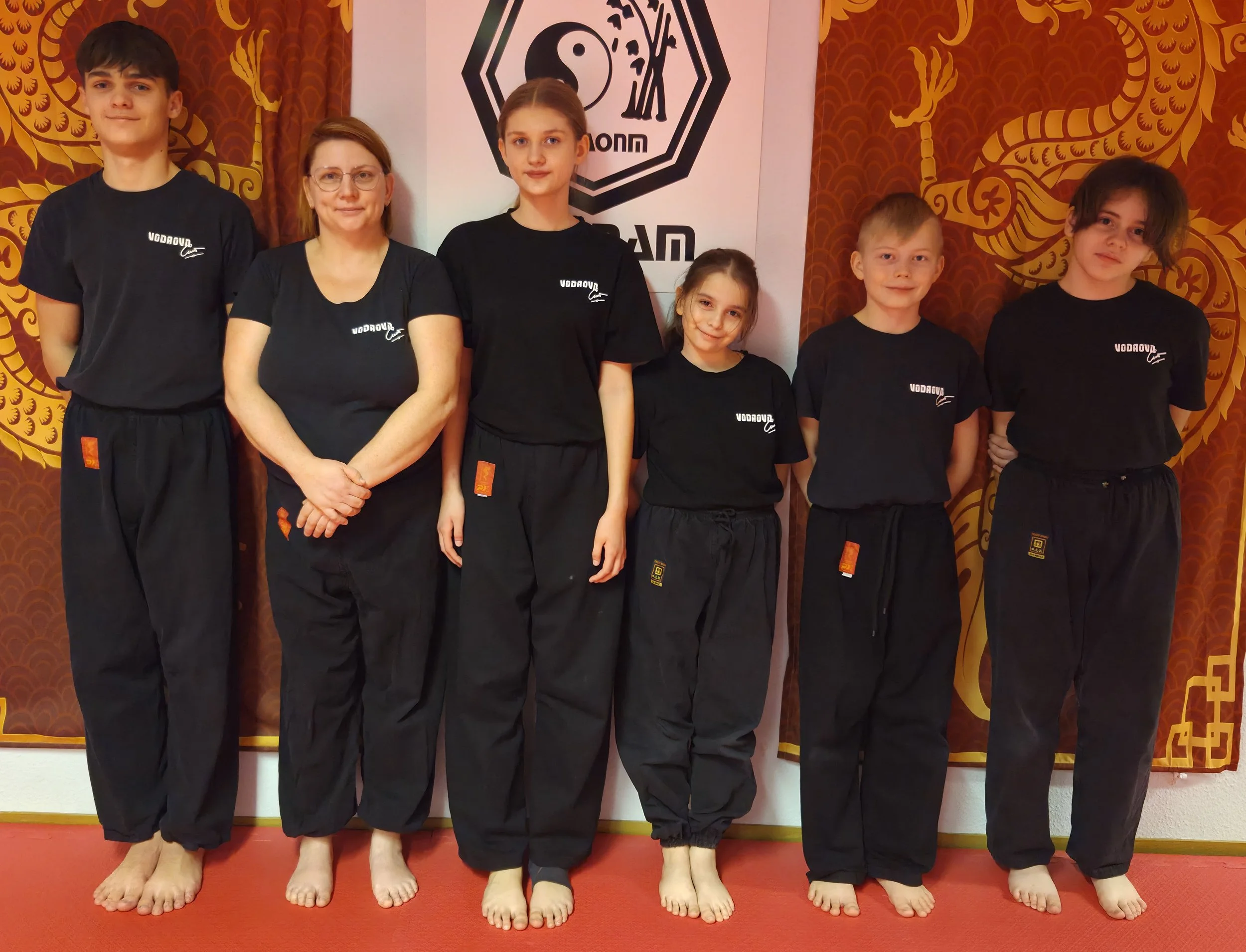 Ein Kinder- und Erwachsenentraining in einem Kampfsportraum mit roten Teppichen und einer Wand mit einem Yin-Yang-Symbol. Sechs Personen stehen in Schulterrückenschau, tragen schwarze Kampfanzüge mit 'VODROUV' auf der Brust.
