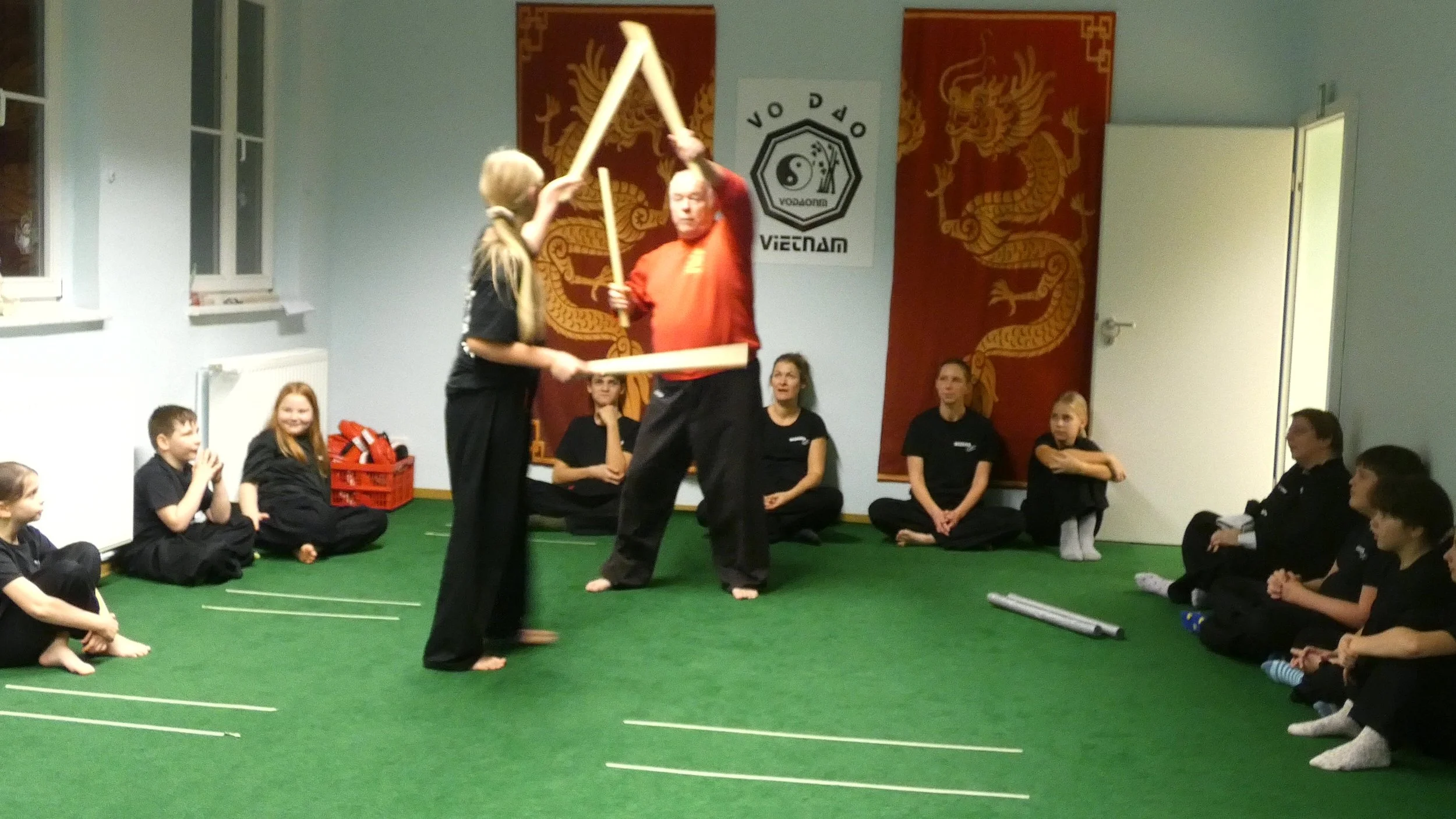 Zwei Martial-Arts-Kämpfer zeigen eine Technik vor einer Gruppe von Kindern und Erwachsenen in einem Kampfsportraum, der mit asiatisch inspirierten Dekorationen verziert ist.