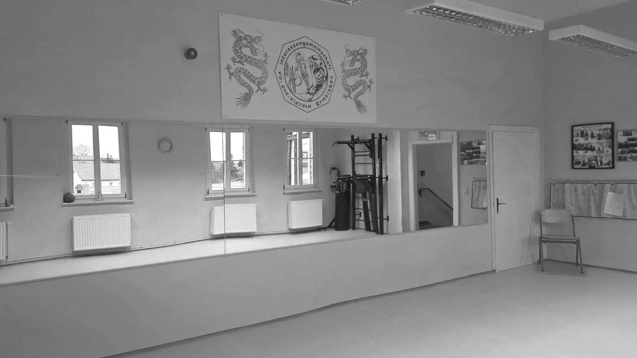 Ein Raum mit mehreren Fenstern, einer Uhr an der Wand, einem Spiegel, einem Stuhl, Fitnessgeräten und einer Tafel mit Fotos an der Wand.