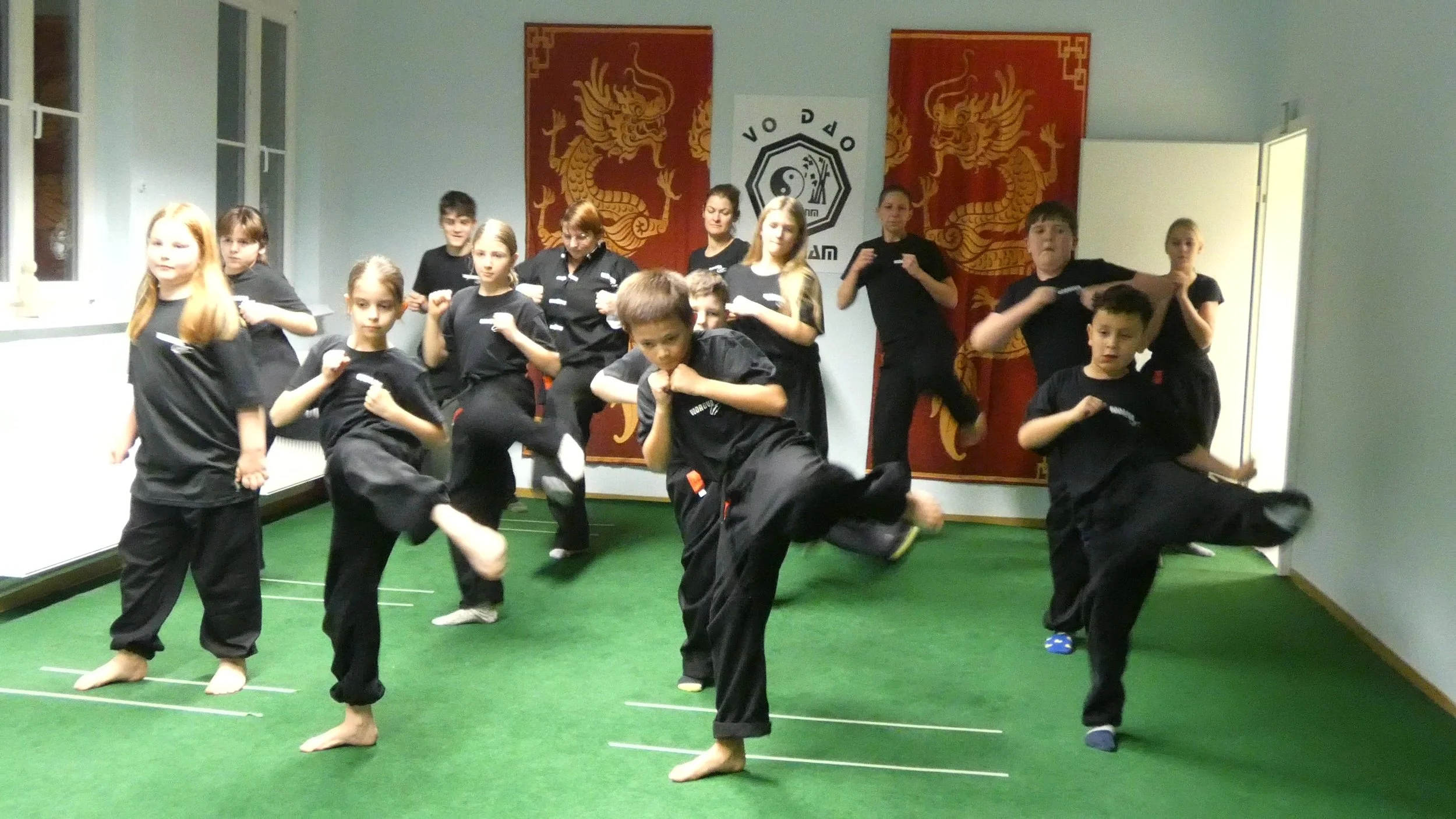 Gruppe von Kindern und Jugendlichen trainieren Taekwondo in einem Raum mit grüner Matte, an der Wand sind chinesische Drachen und ein Yin-Yang-Symbol. Alle sind in schwarzen Taekwondo-Gis gekleidet.