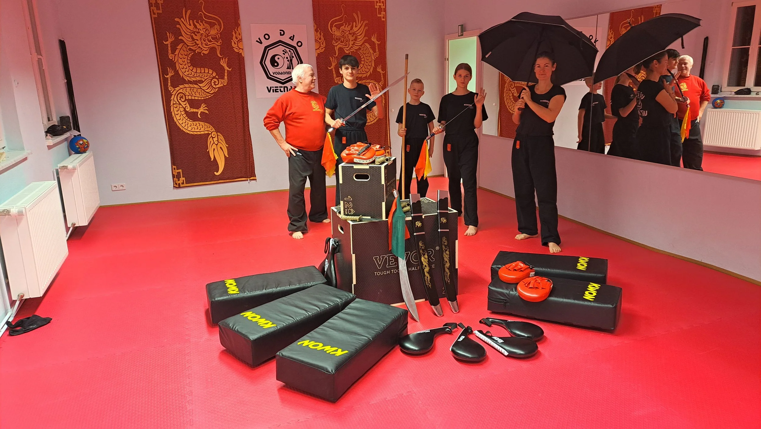 Ein Gruppenfoto in einer Kampfsportschule mit mehreren Personen, die Kampfsportausrüstung und -trainingsequipment tragen, vor einem Wandbild mit einem chinesischen Drachen und einem Yin-Yang-Symbol.