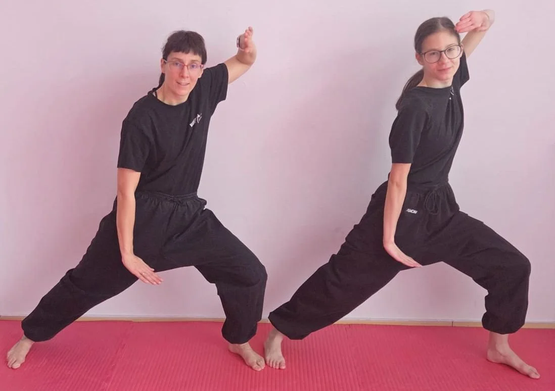 Zwei Frauen in schwarzen Sportkleidung, die eine Kampfsportposition im Makhi verschaffen, stehen auf einer roten Matte vor einer weißen Wand.