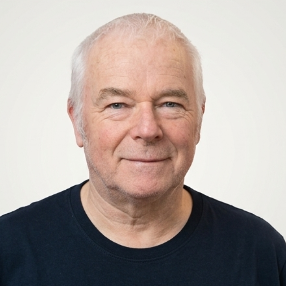 Porträt eines älteren Mannes mit weißen Haaren, blauen Augen und einem schwarzen T-Shirt, lächelnd vor einem weißen Hintergrund.