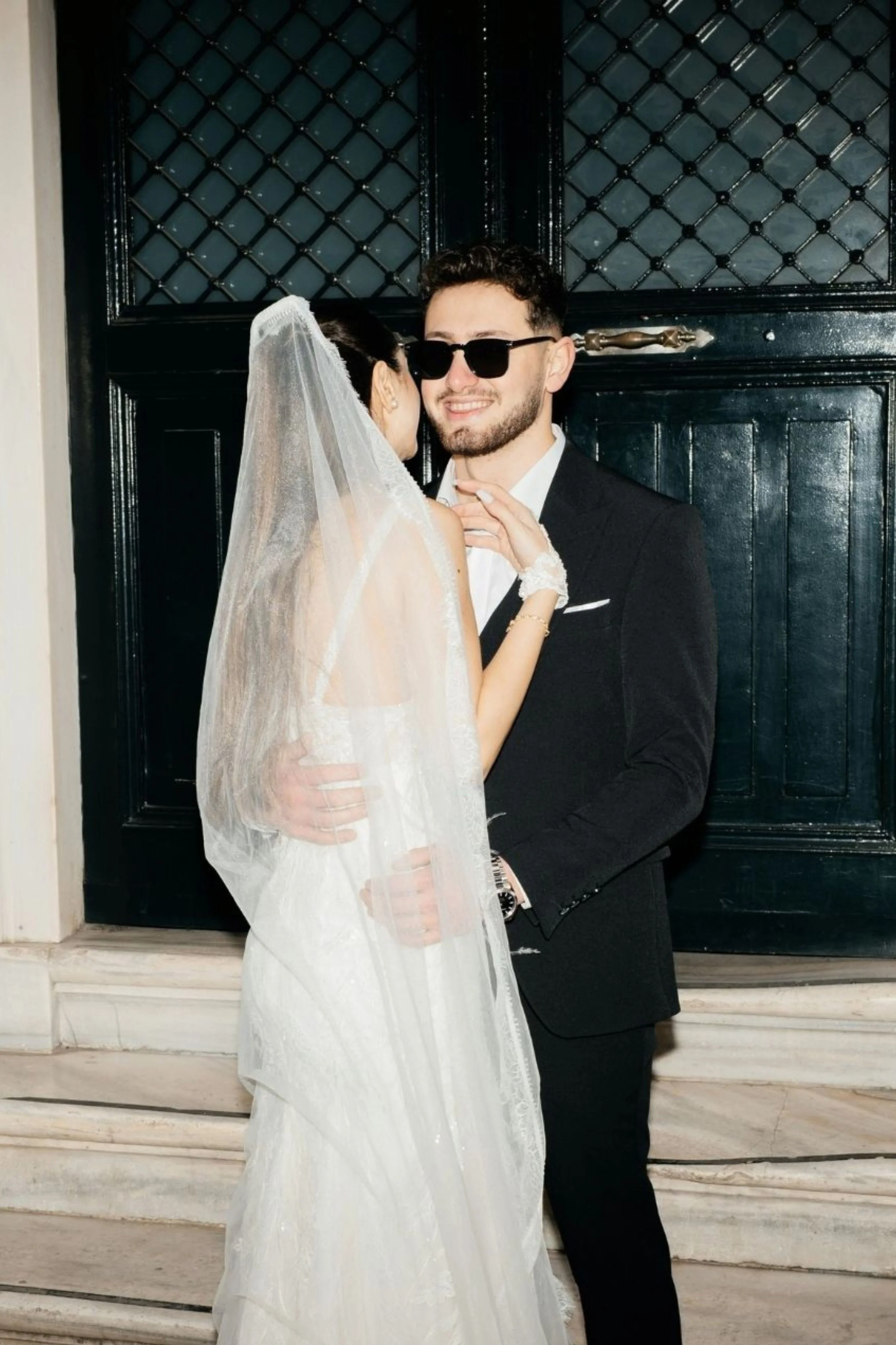 A couple dressed in wedding attire standing in front of a black door. The woman is wearing a wedding dress and veil, and the man is wearing a black suit with sunglasses.