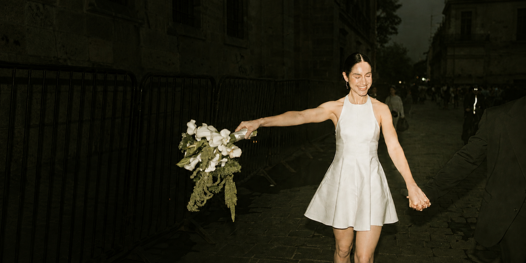 A woman in a white dress holding a bouquet of white flowers smiling while holding hands with a person in a black suit, outdoors at night.
