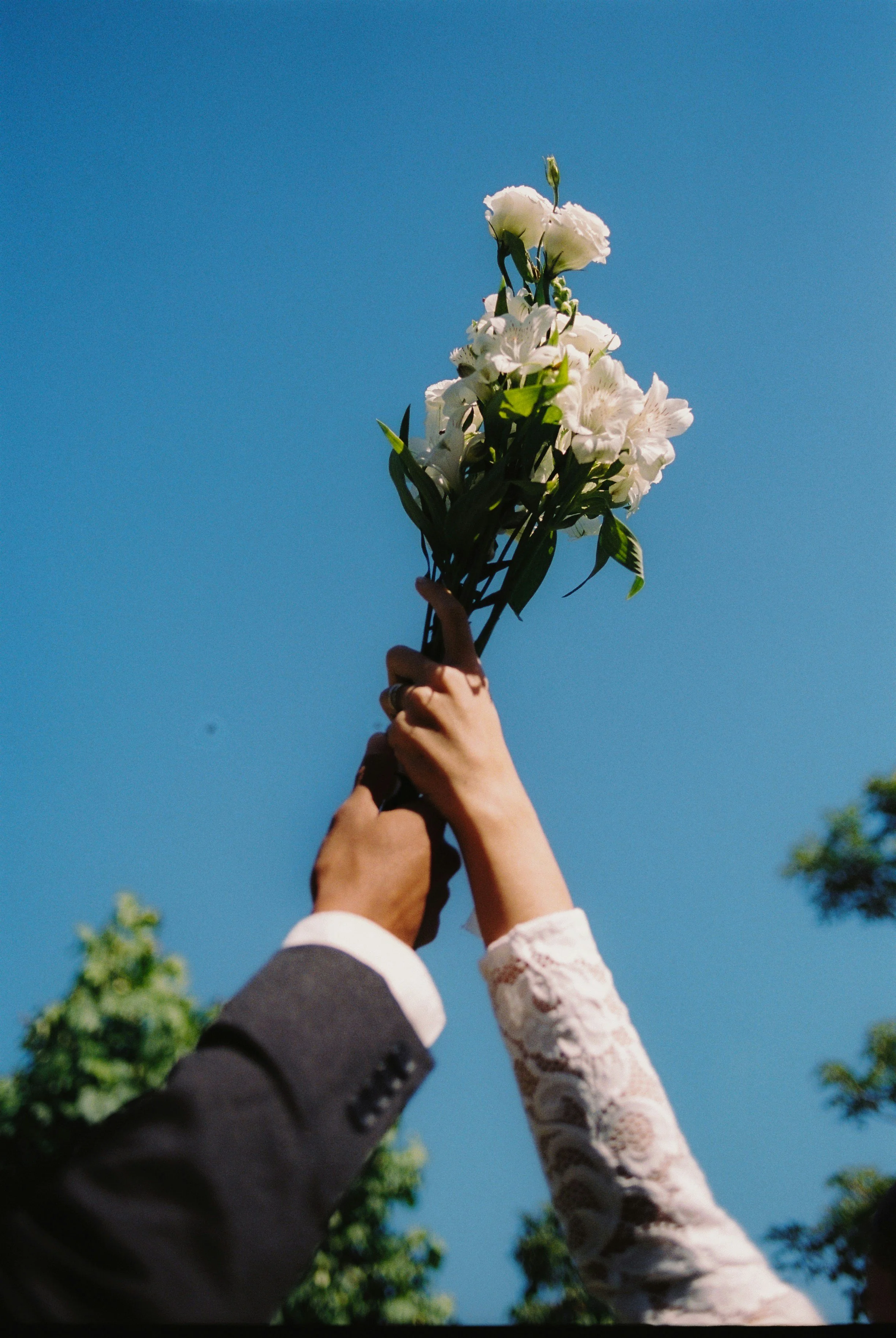 Two people holding a bouquet of white flowers up to the clear blue sky.