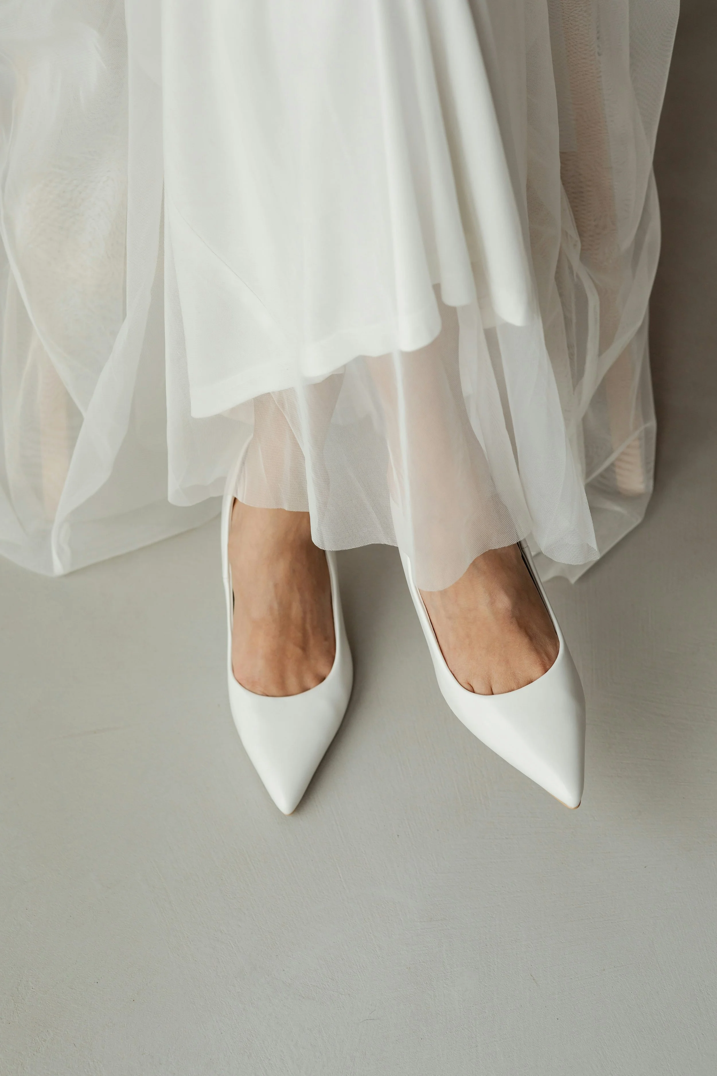 Close-up of a woman in a white wedding dress with sheer, layered fabric, wearing white high-heeled shoes with pointed toes, standing on a light-colored floor.