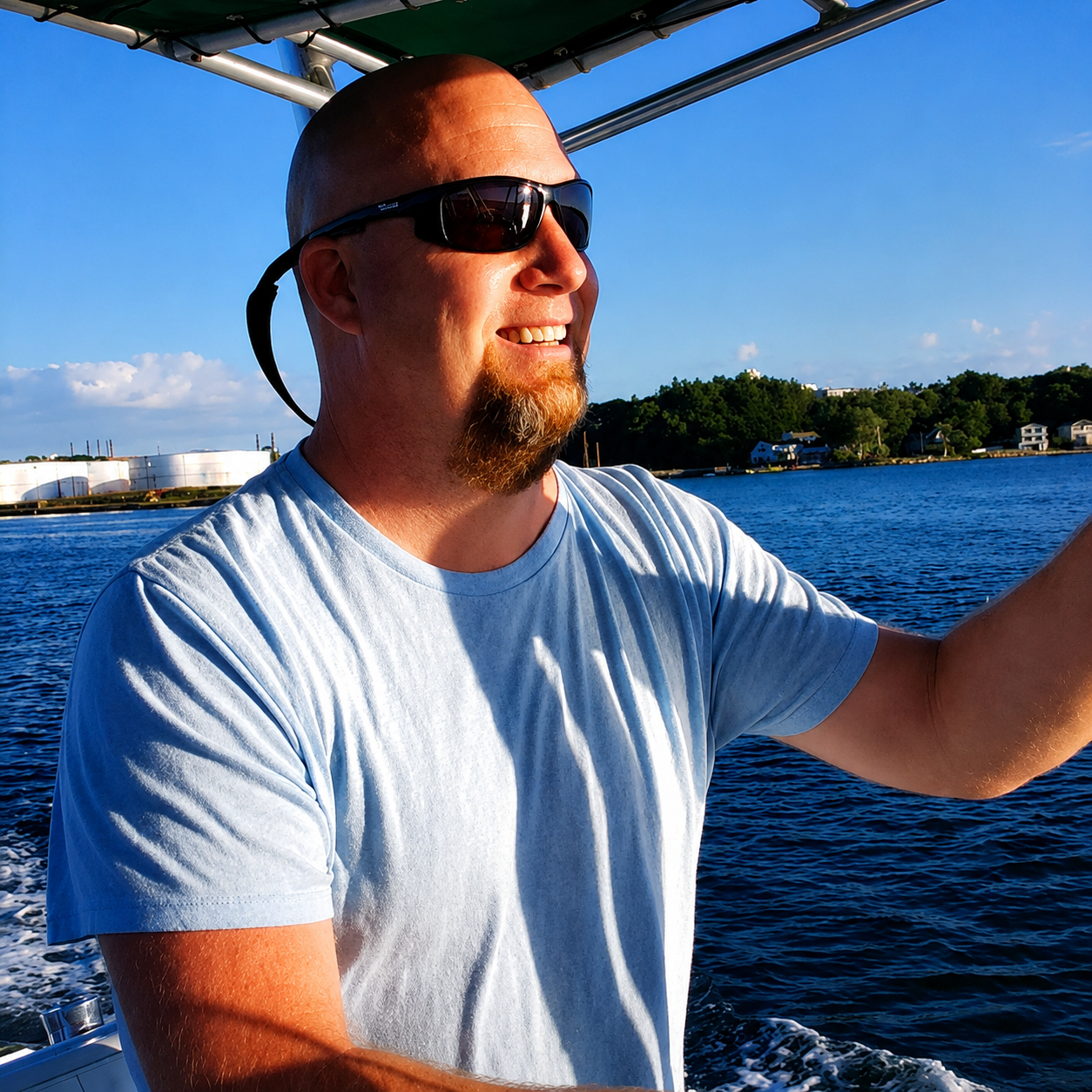A man with sunglasses and a beard smiling while on a boat on the water during sunset, with a shoreline, houses, and trees in the background.