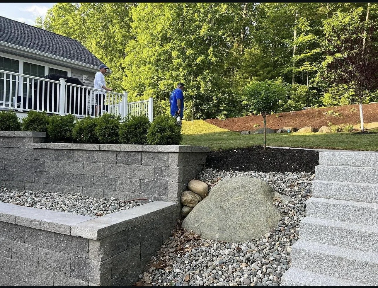 A landscaped backyard with stone retaining walls, gravel, a small tree, and two people standing on a deck overseeing the garden.