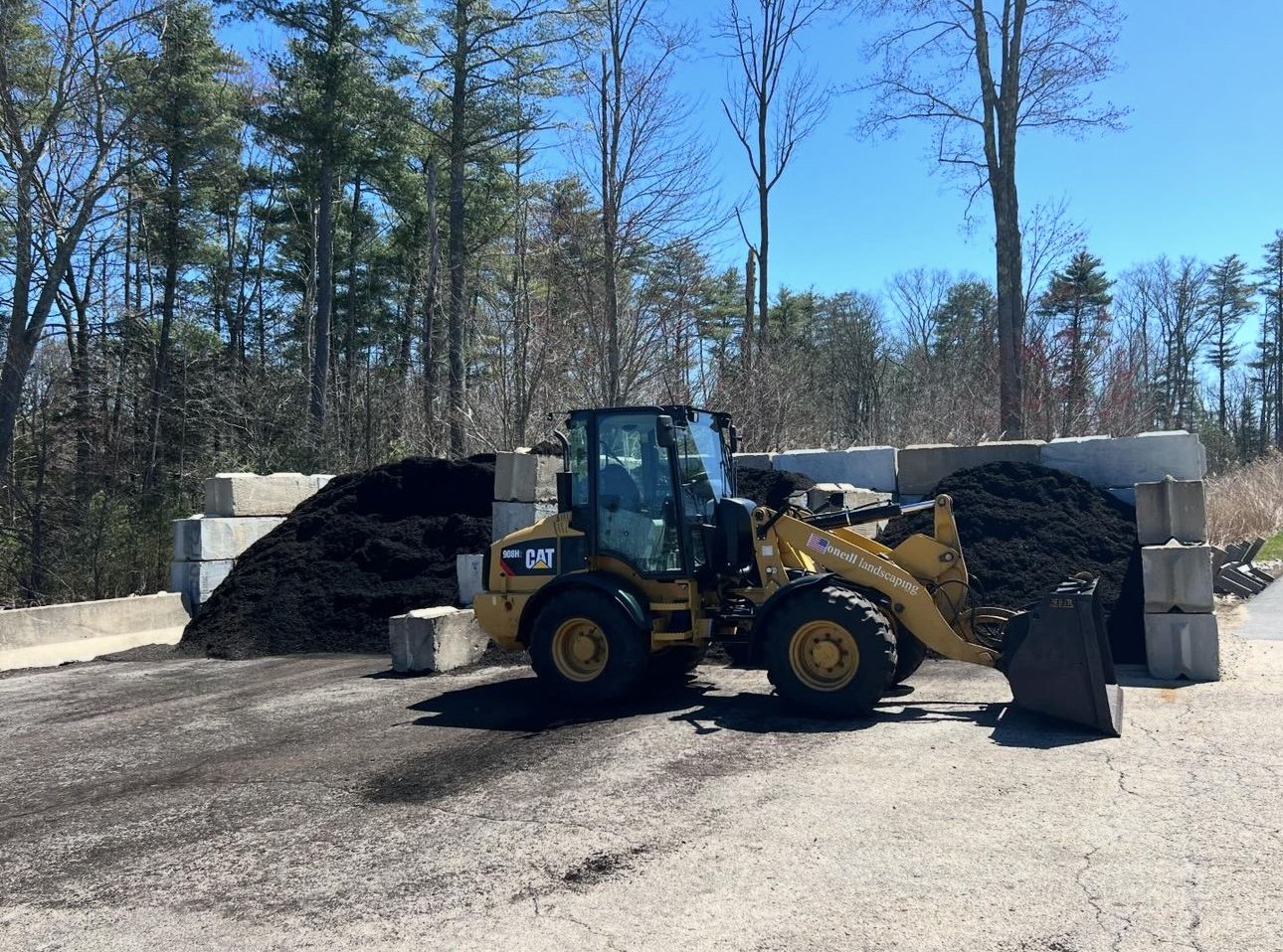 A front-end loader parked on a paved area with large piles of dark soil or mulch behind it. There are concrete barriers along the edge and a forest of tall trees with some bare branches in the background under a clear blue sky.