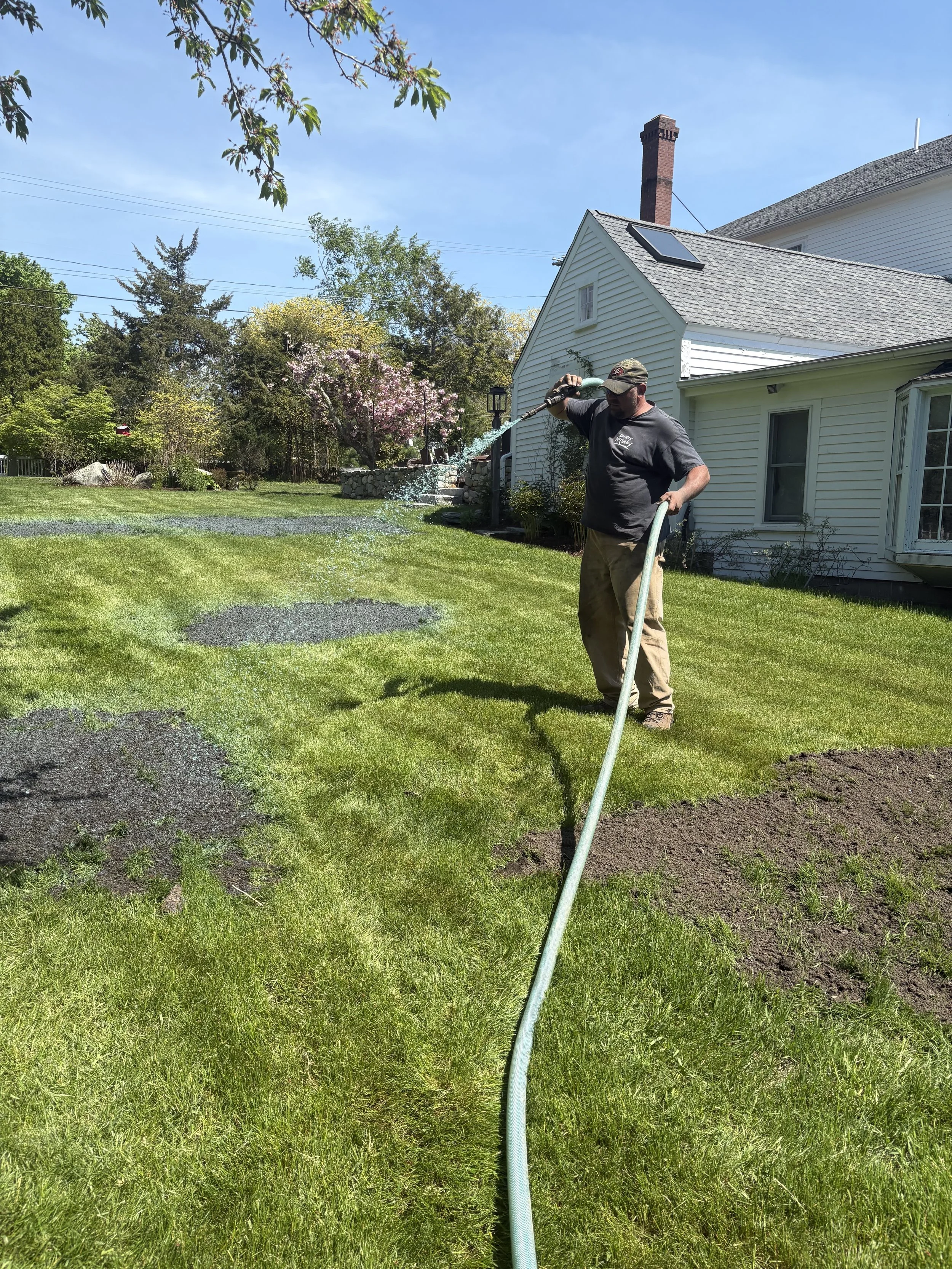 A man watering a lawn with a garden hose in a backyard of a white house on a sunny day.