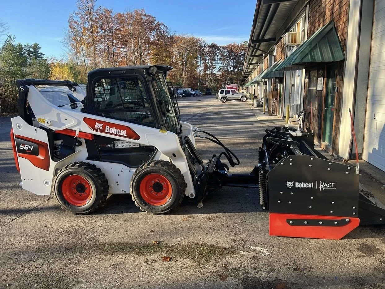 A Bobcat skid-steer loader with a KAGE blower attachment parked outside a building with green awnings, with trees and parked cars in the background.