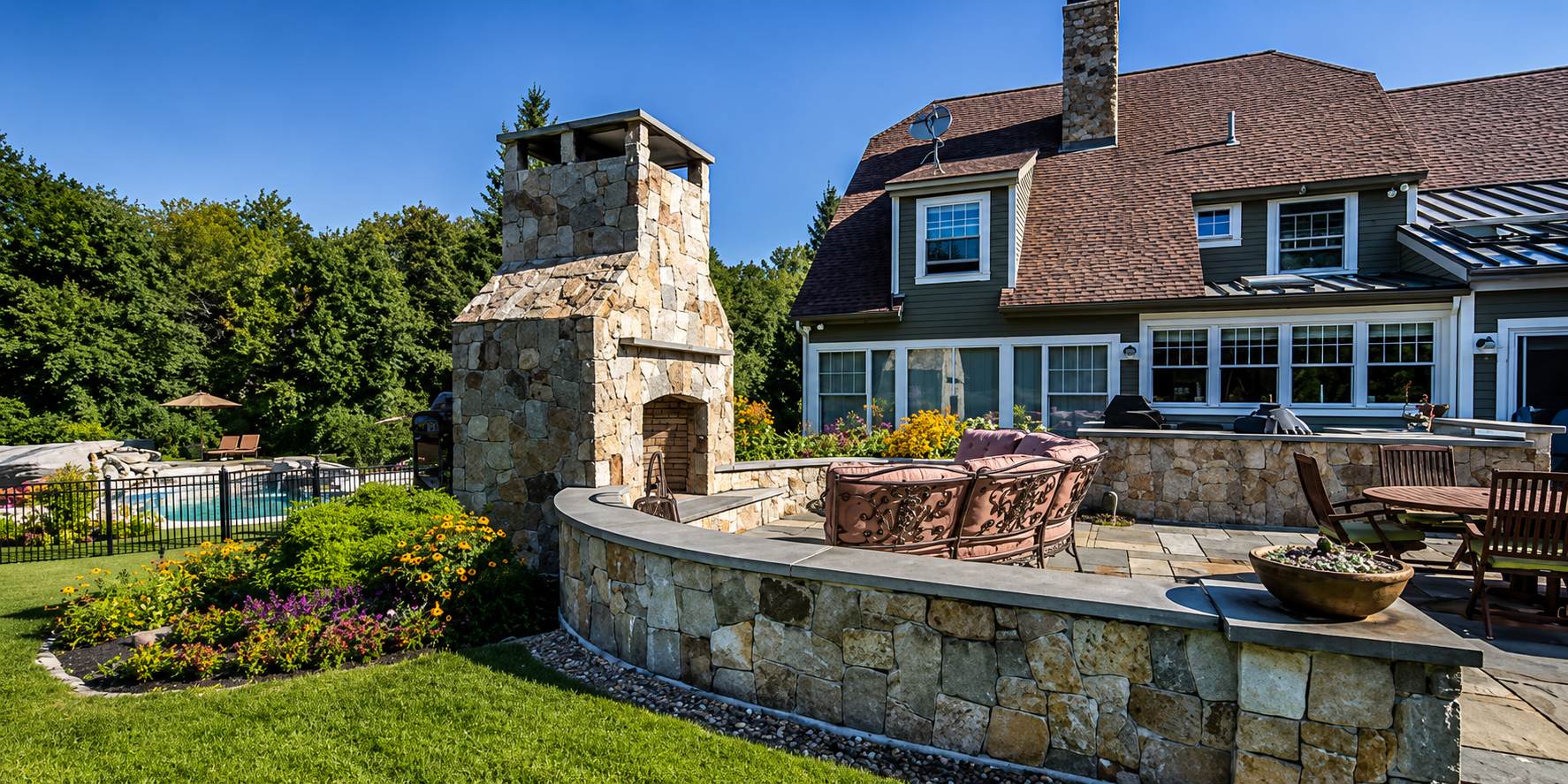 Backyard with stone outdoor fireplace, patio furniture, swimming pool, and garden, adjacent to a house with large windows and a brown shingle roof, surrounded by green trees.