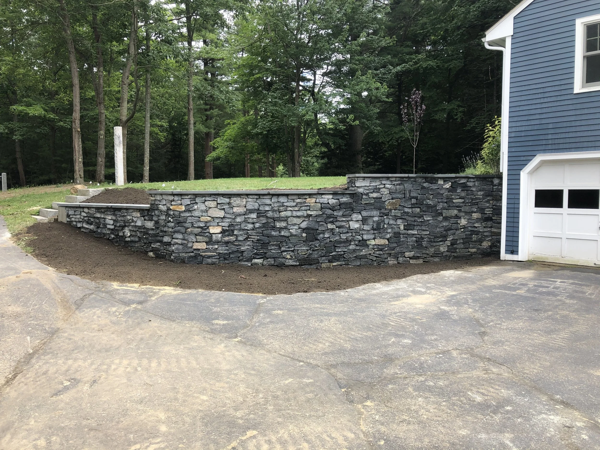 A retaining wall made of stacked flat stones, with a grassy area and trees behind it. Part of a blue house with white trim and a garage door on the right side.