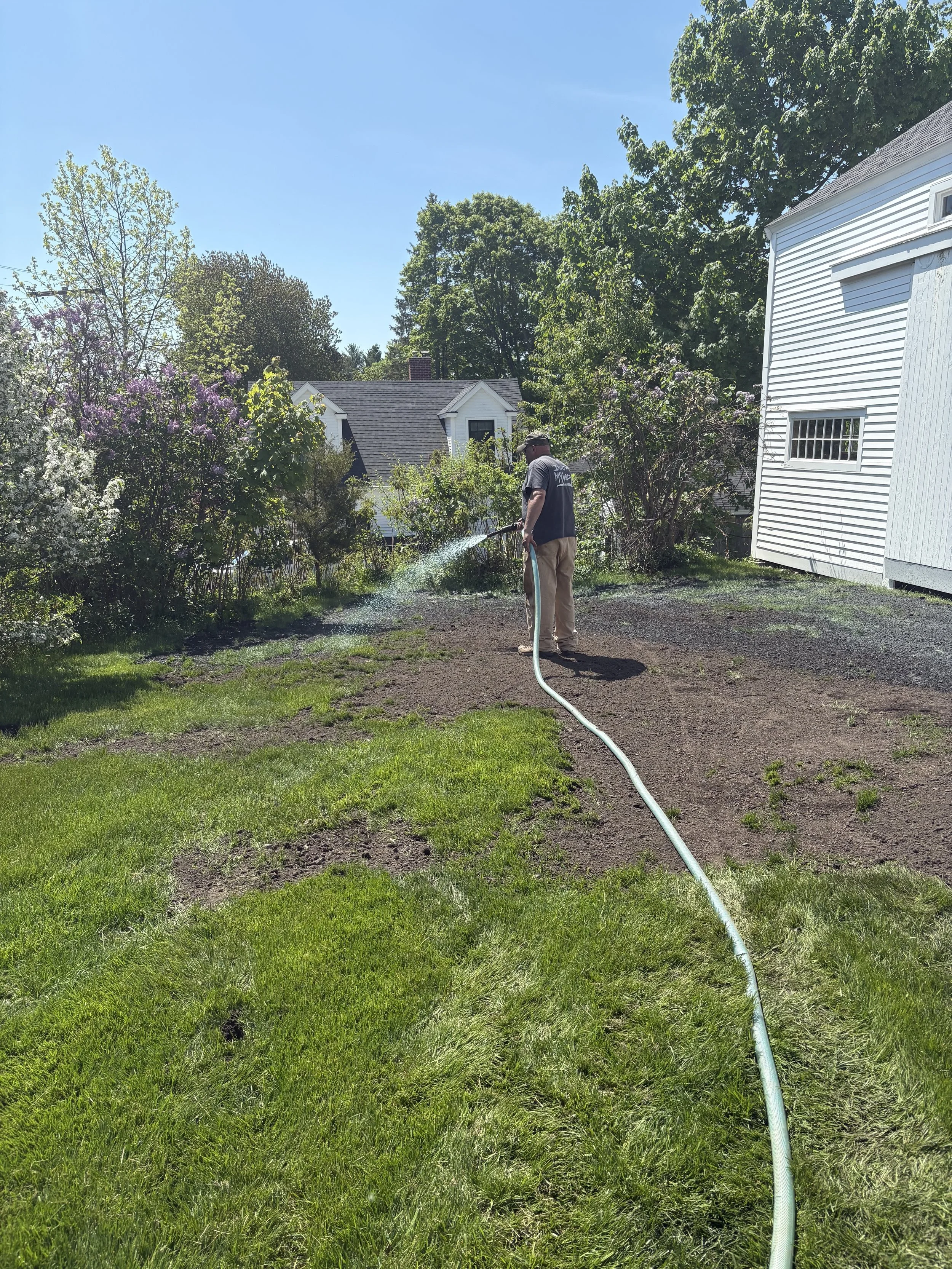 A man watering a garden with a garden hose next to a white house on a sunny day with clear blue sky and green trees.