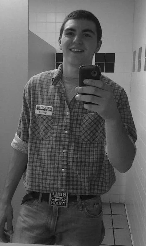 A young man taking a selfie in a public restroom, wearing a plaid shirt with a name tag and jeans.