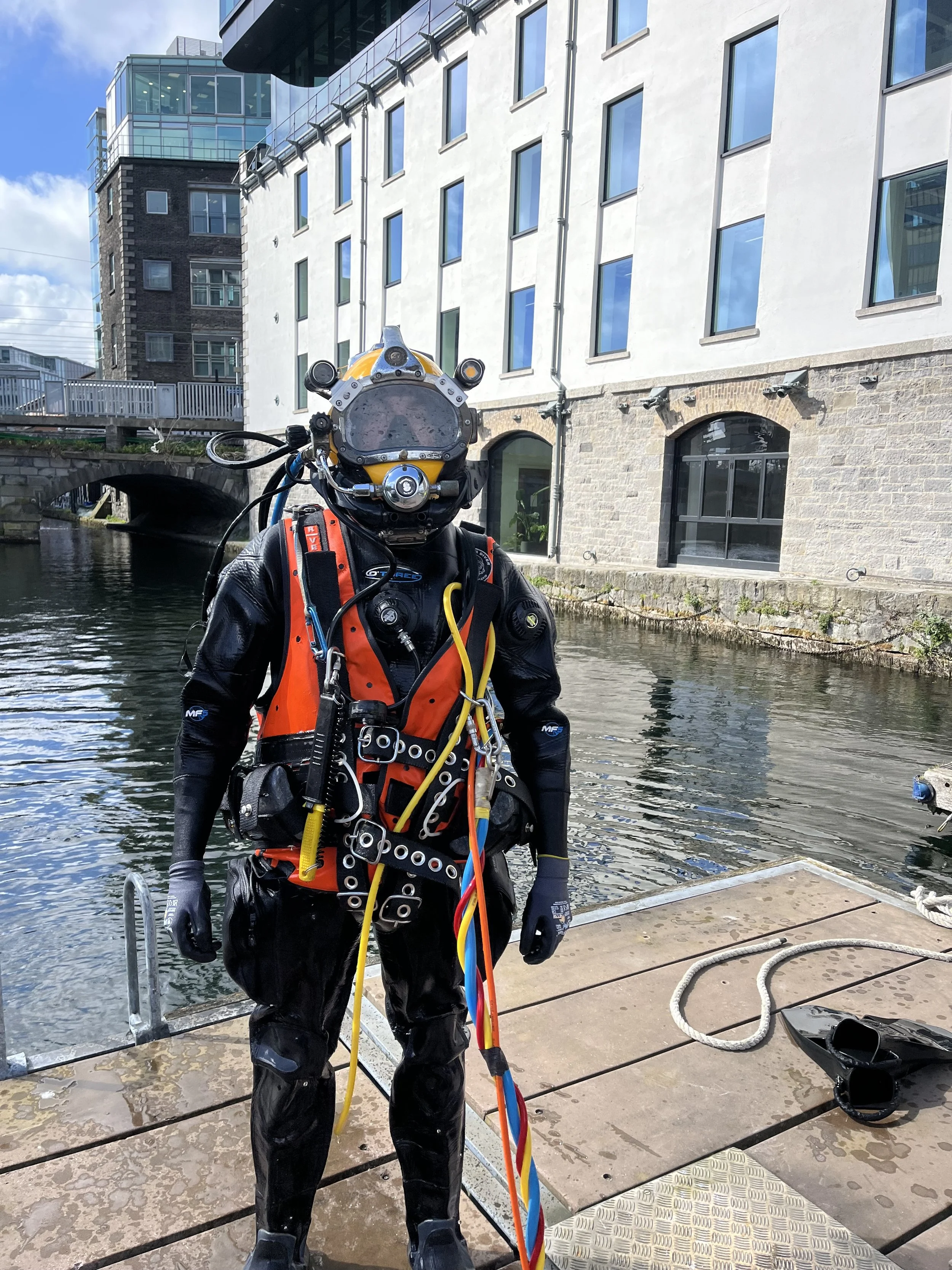 A diver in a wetsuit standing on a dock near the water, with a modern white building with large windows and a brick facade in the background.
