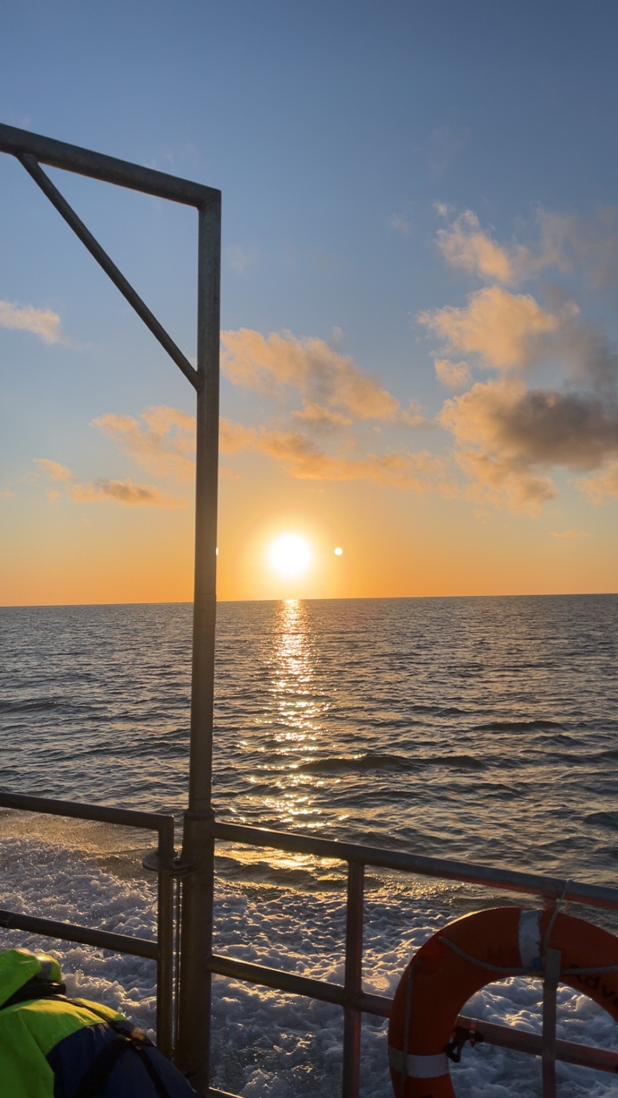 A sunset over the ocean as seen from a boat, with metal guardrails and a life preserver on the boat.