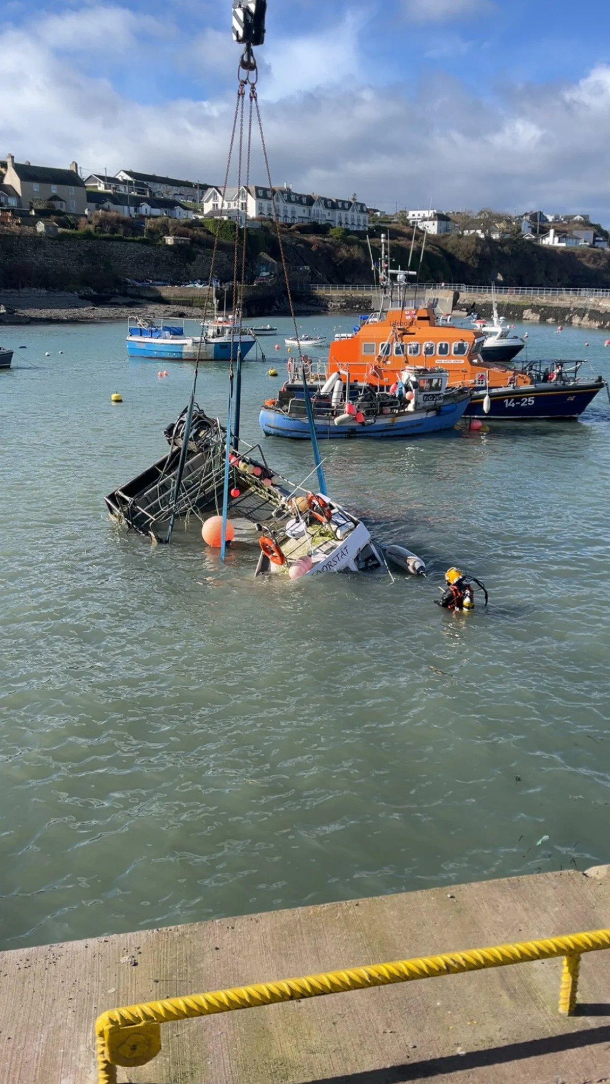 A boat is sinking in a harbor with emergency responders attending to the scene, and a crane is lifting the boat out of the water.