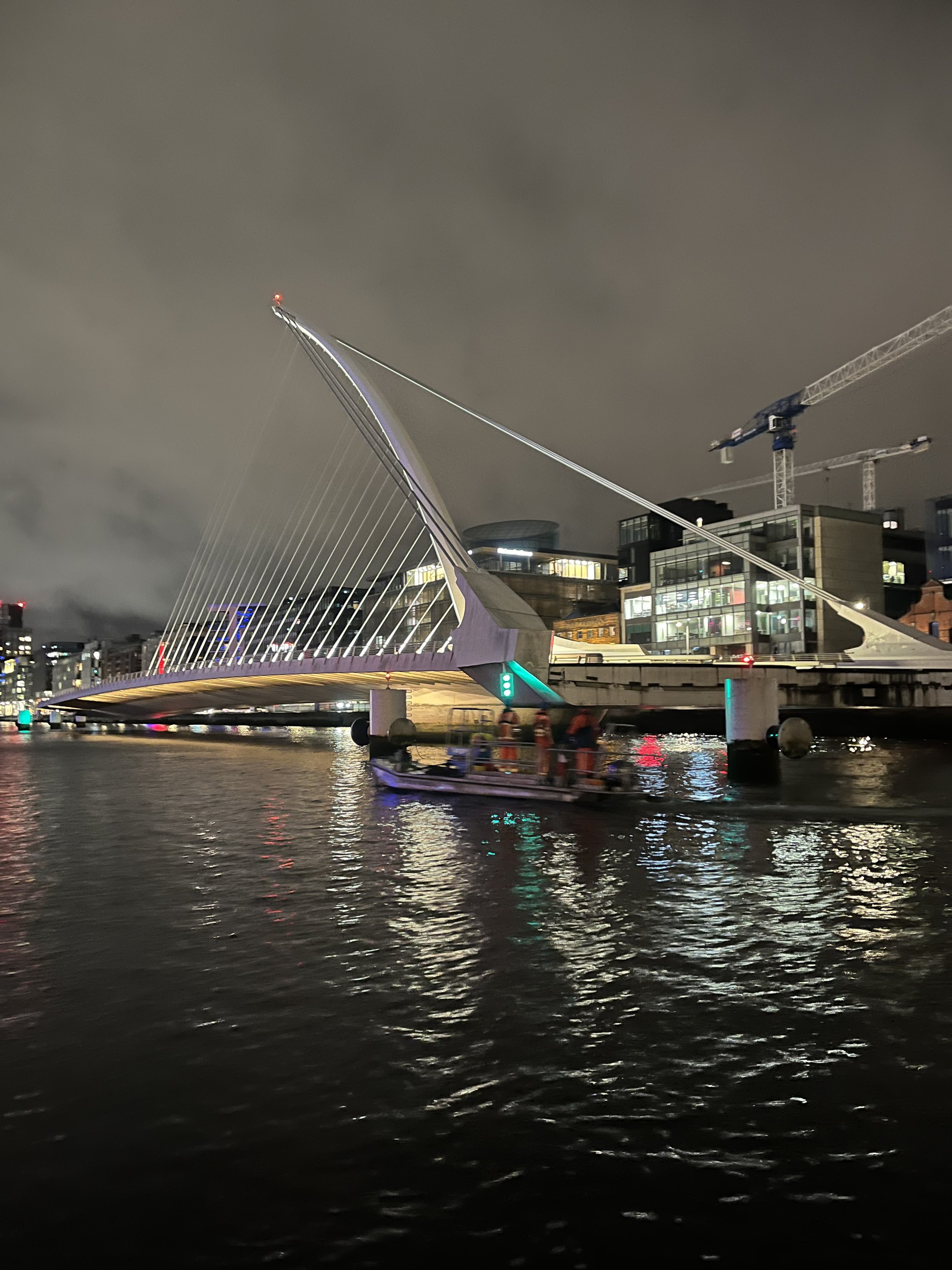 Nighttime cityscape with a modern suspension bridge illuminated, a boat on water reflecting the lights, and construction cranes in the background.