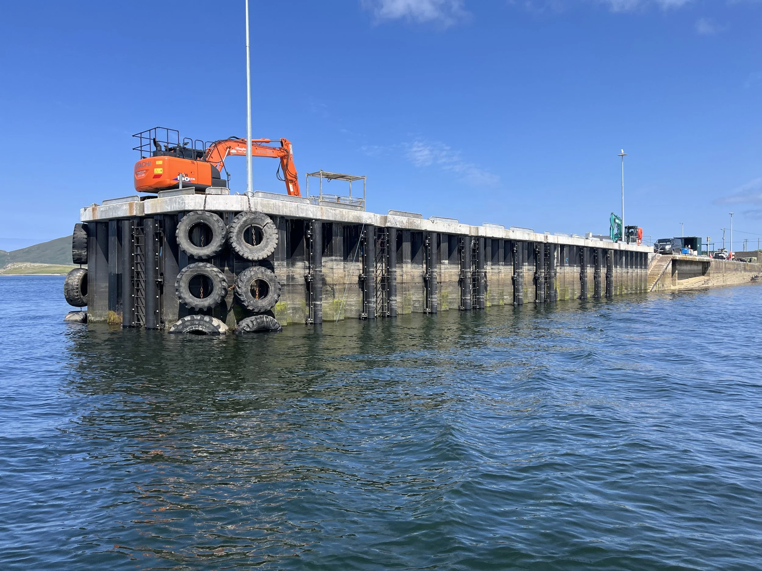 A large dock extends over water with a small orange excavator on it, and lots of tires hanging on its sides, with a blue sky above.
