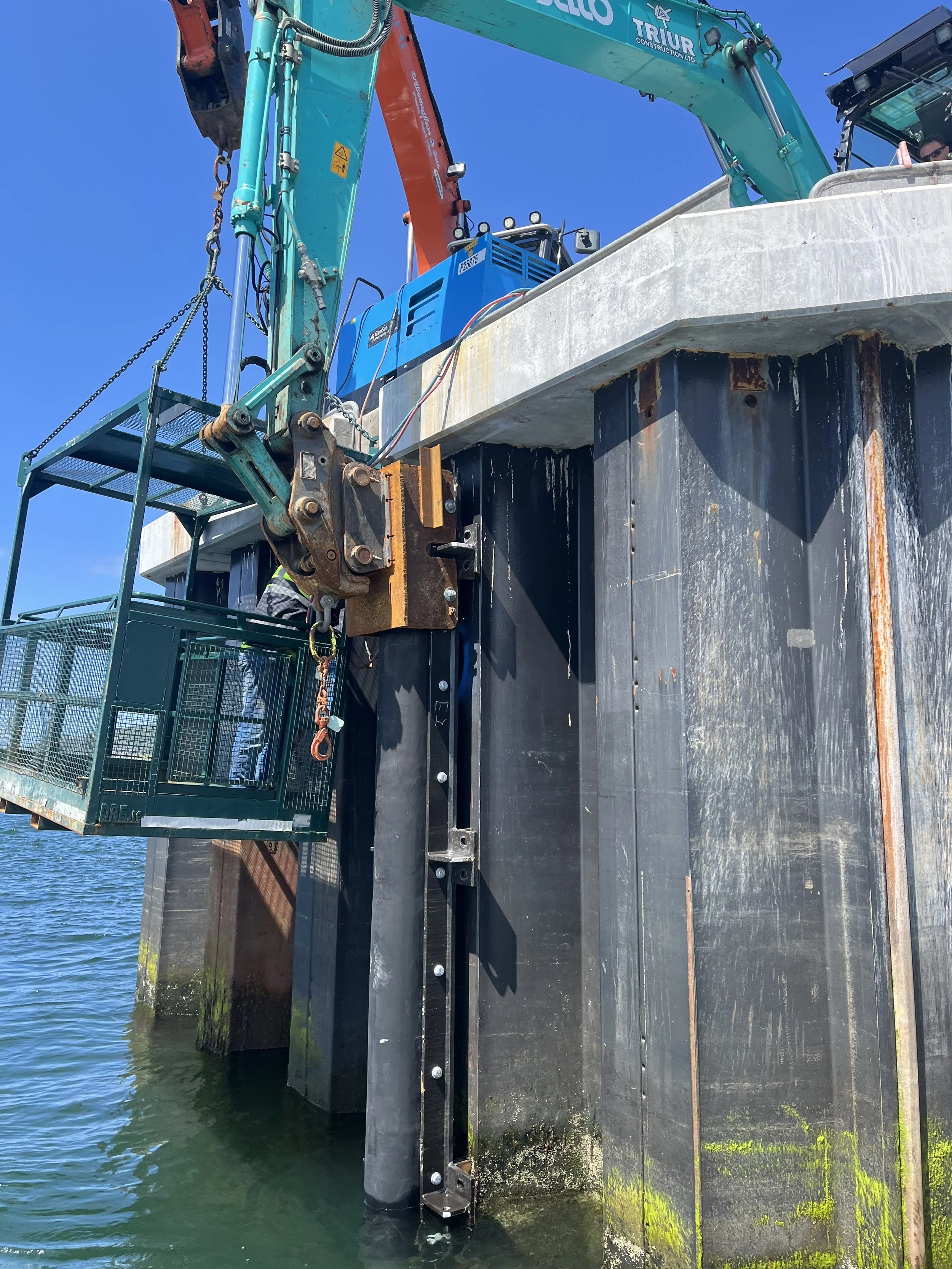 Construction workers using heavy machinery to build a structure over water, with a large excavator and safety cage under a clear blue sky.