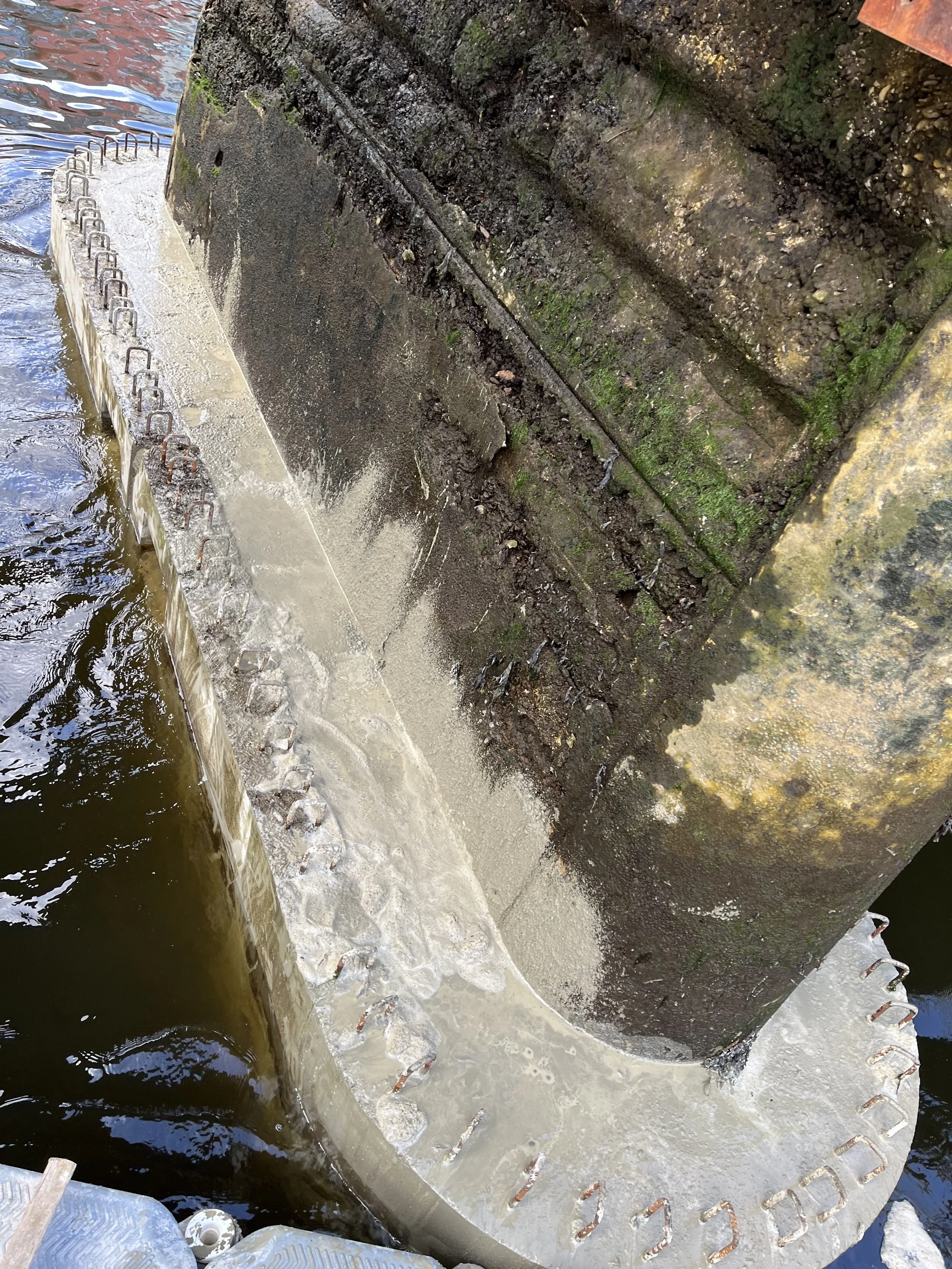 A large concrete and metal structure with water flowing over the edge, possibly a dam or water intake structure, with rusted metal hooks and green algae on the surface.