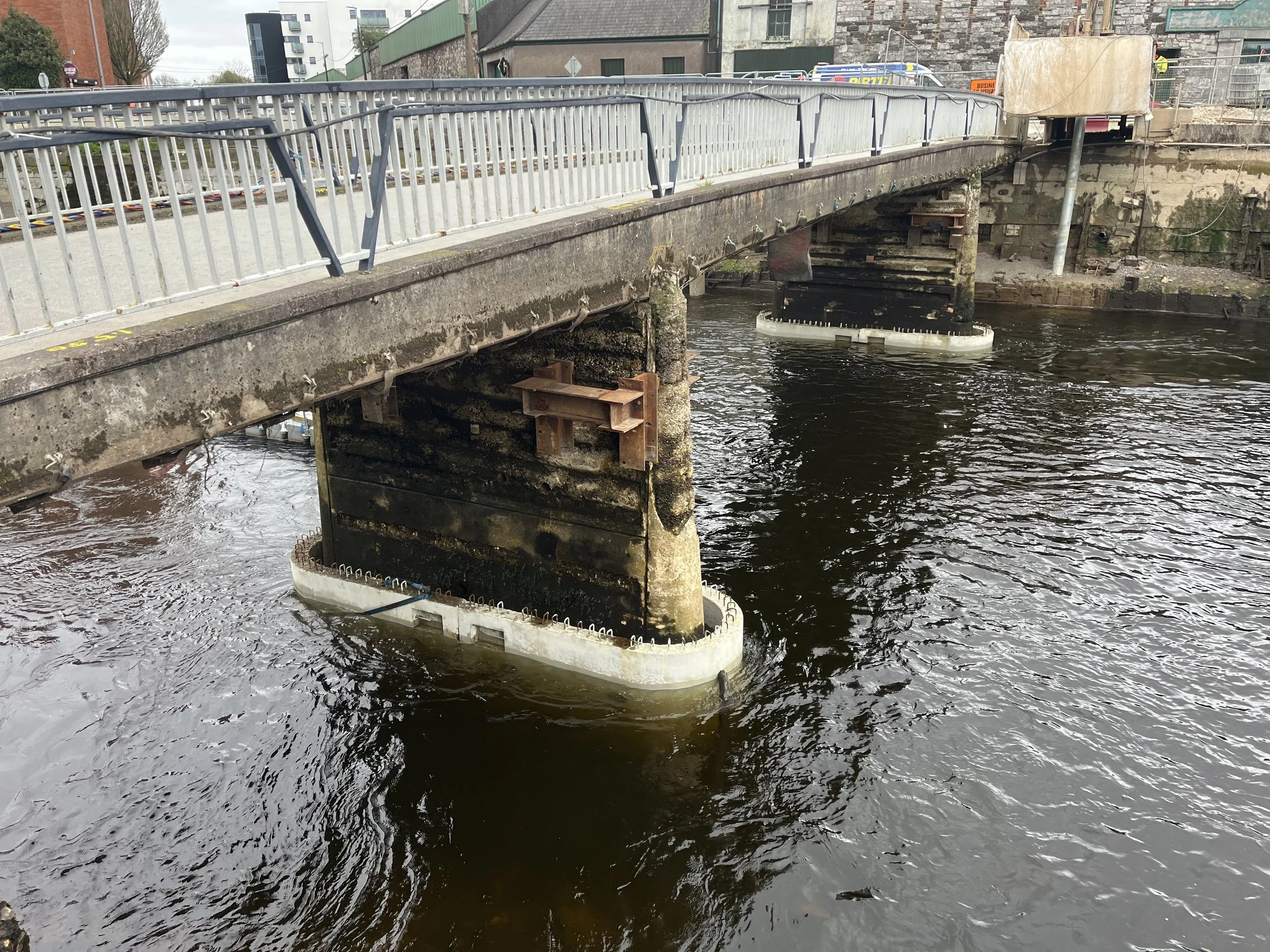 A bridge with a metal railing over a body of water, with the bridge undergoing construction or repair, as indicated by the temporary guardrail and construction sign in the background.