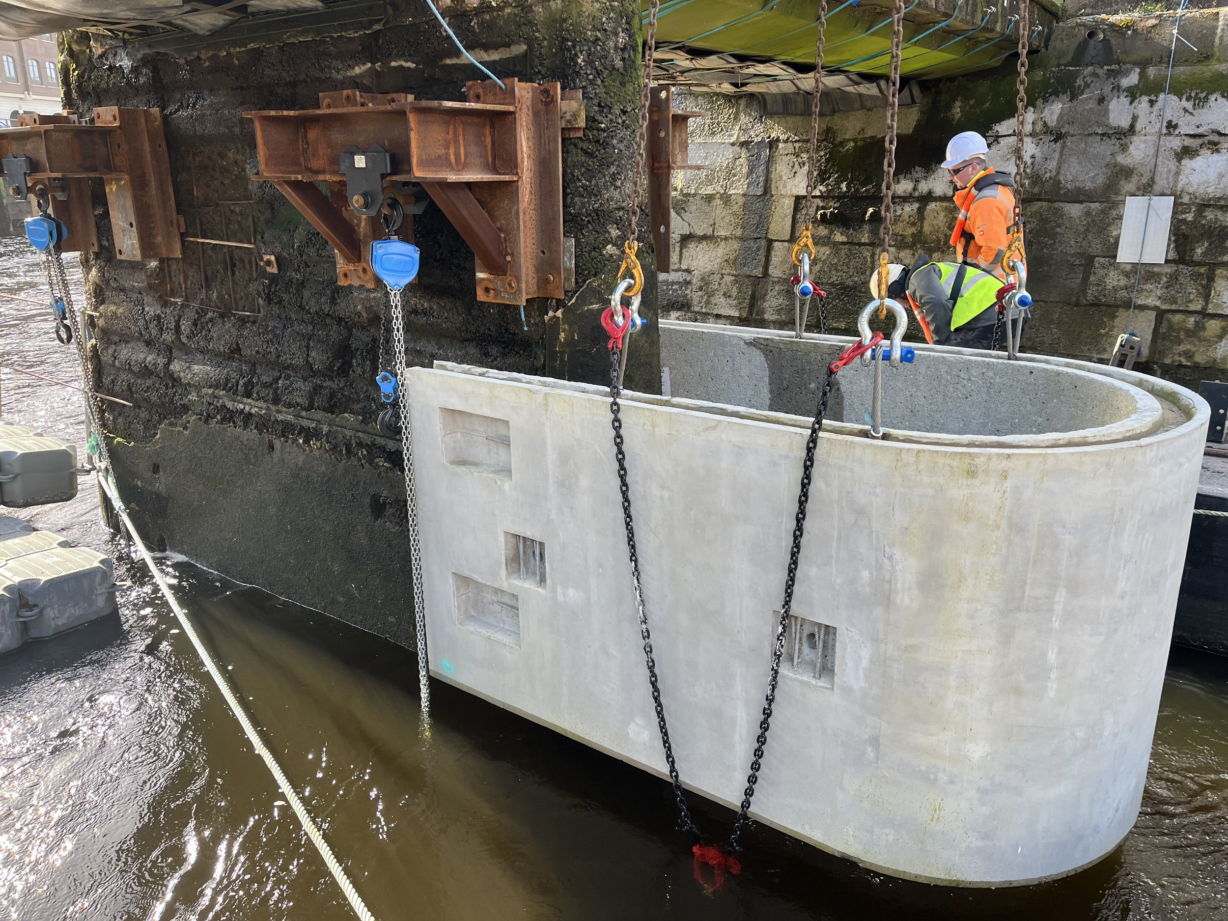 Construction workers installing a large concrete structure in water, supported by rusted steel beams and chains, with safety gear including helmets and reflective vests.
