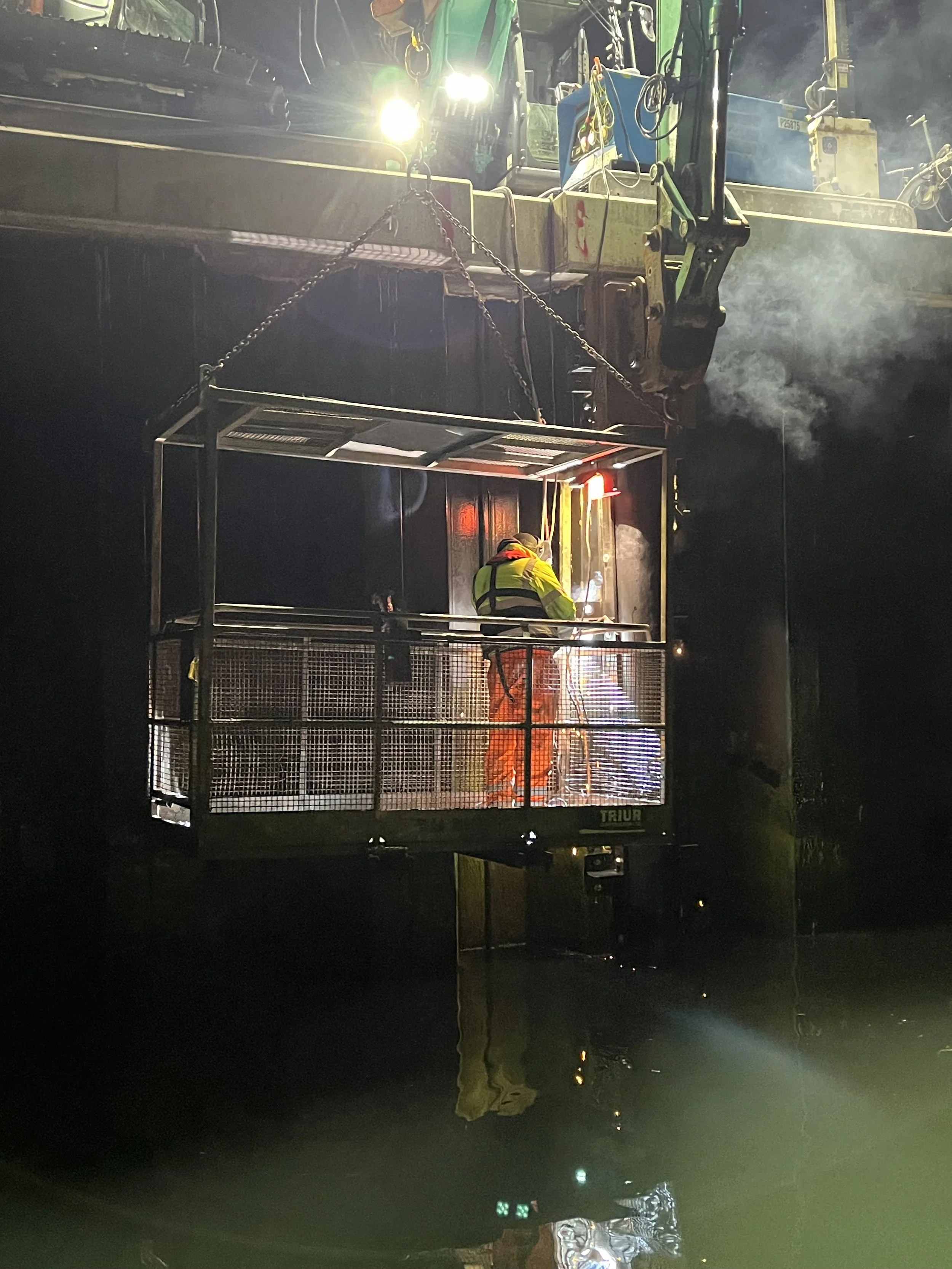 A construction worker wearing a high-visibility vest and gloves is on a platform, welding or working with metal, while a large crane supports the platform. The scene appears to be during nighttime.