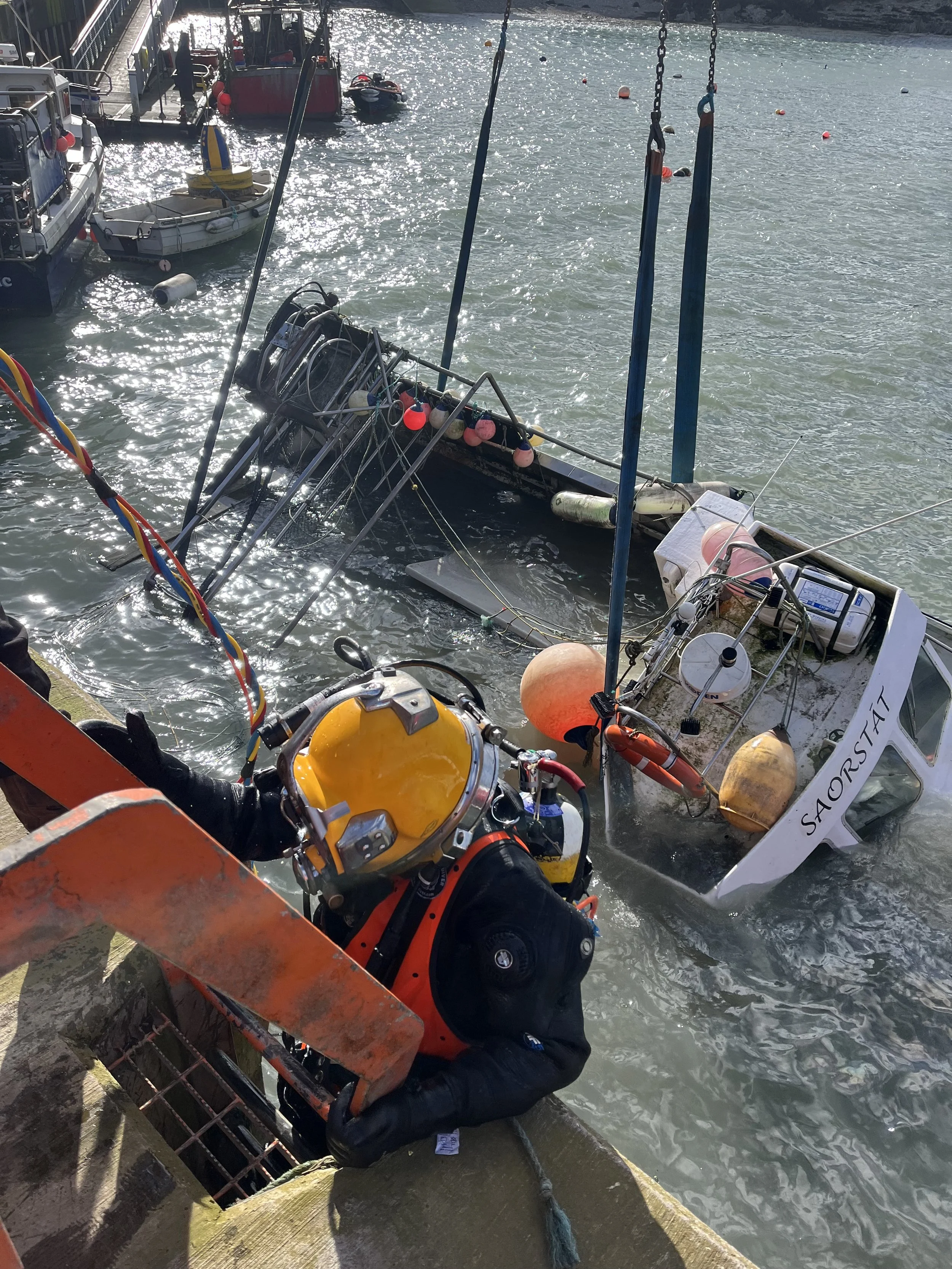 Rescue worker in black wetsuit and orange life vest onboard a partially submerged boat performing a rescue operation during daylight at the dock, with boats and water in the background.