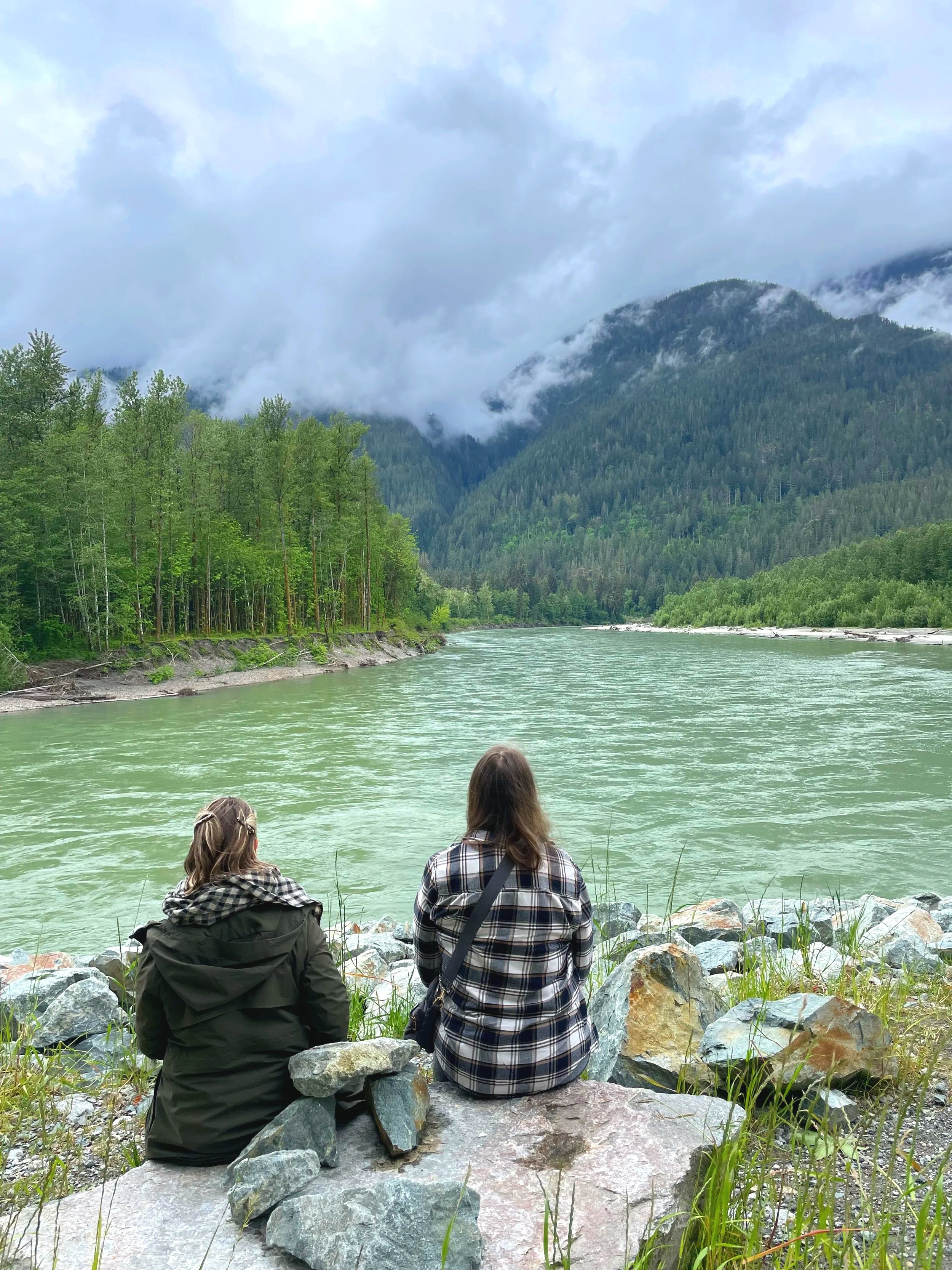 Two women sitting by a river, surrounded by rocks, with forested mountains and cloudy sky in the background.